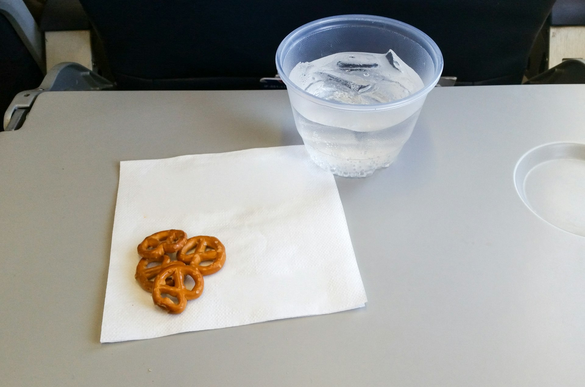 A small pile of pretzels rest on the corner of a white napkin on an airline tray. Next to the napkin is plastic cup filled with water and ice.