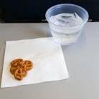 A small pile of pretzels rest on the corner of a white napkin on an airline tray. Next to the napkin is plastic cup filled with water and ice.