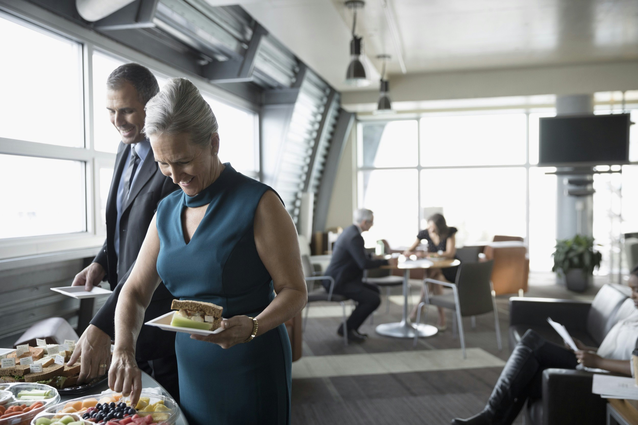 A man and woman pick up food from a buffet table as other travelers sit in comfortable chairs in an airport lounge; Upgrade your travel game