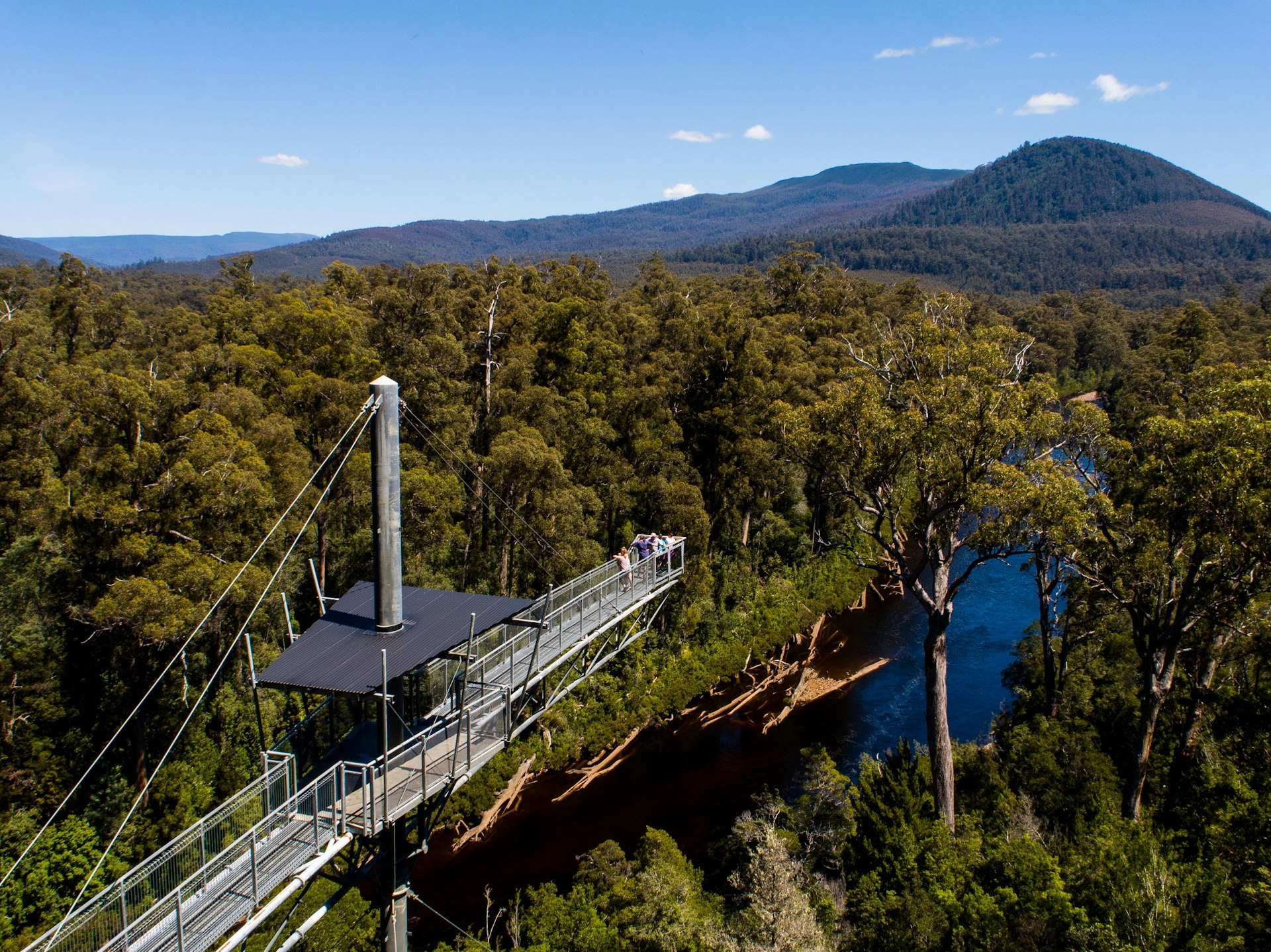 Shot from above, the image looks down on a metal walkway that cantilevers over a section of river; the whole scene is dominated by the forest, which extends into the distant hills.