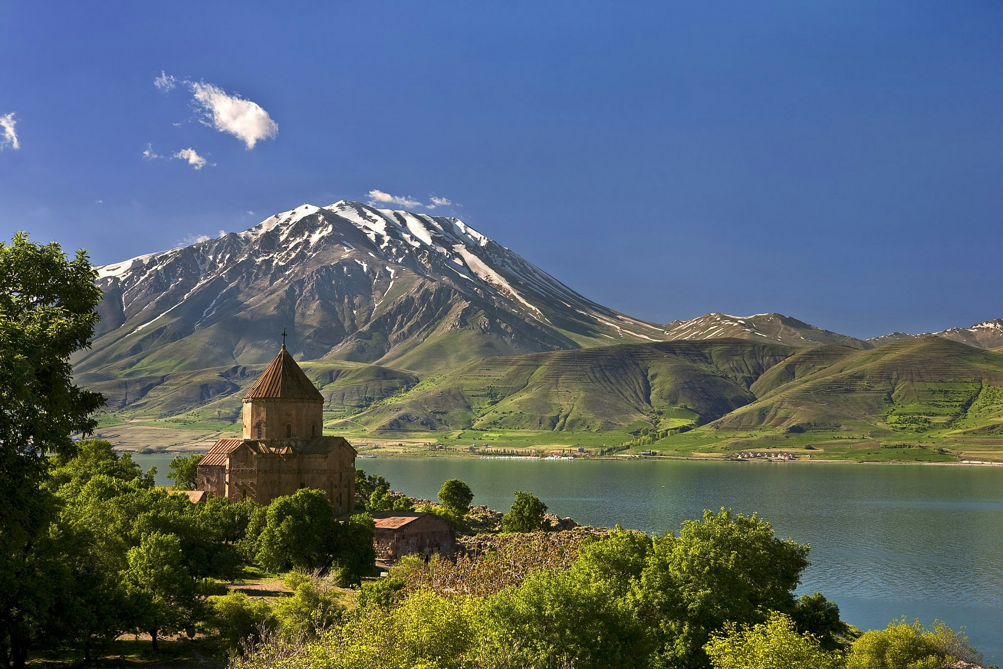 A conical domed church on an island in a lake, with a huge snow-capped mountain rising up from the shore beyond.