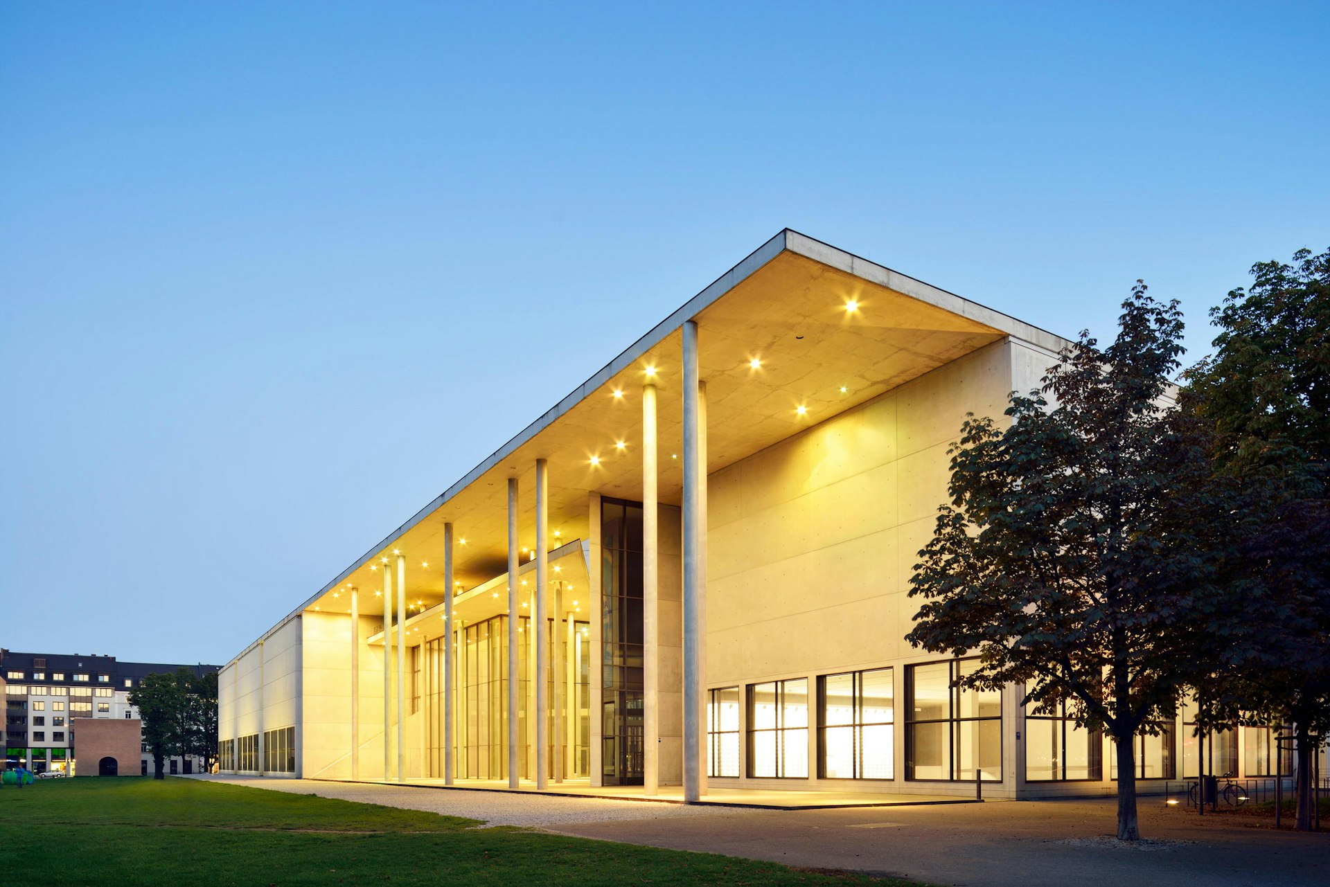 Dusk shot of a vast white modern building with large square windows and thin columns supporting the roof. There are a couple of trees to the left of the building.