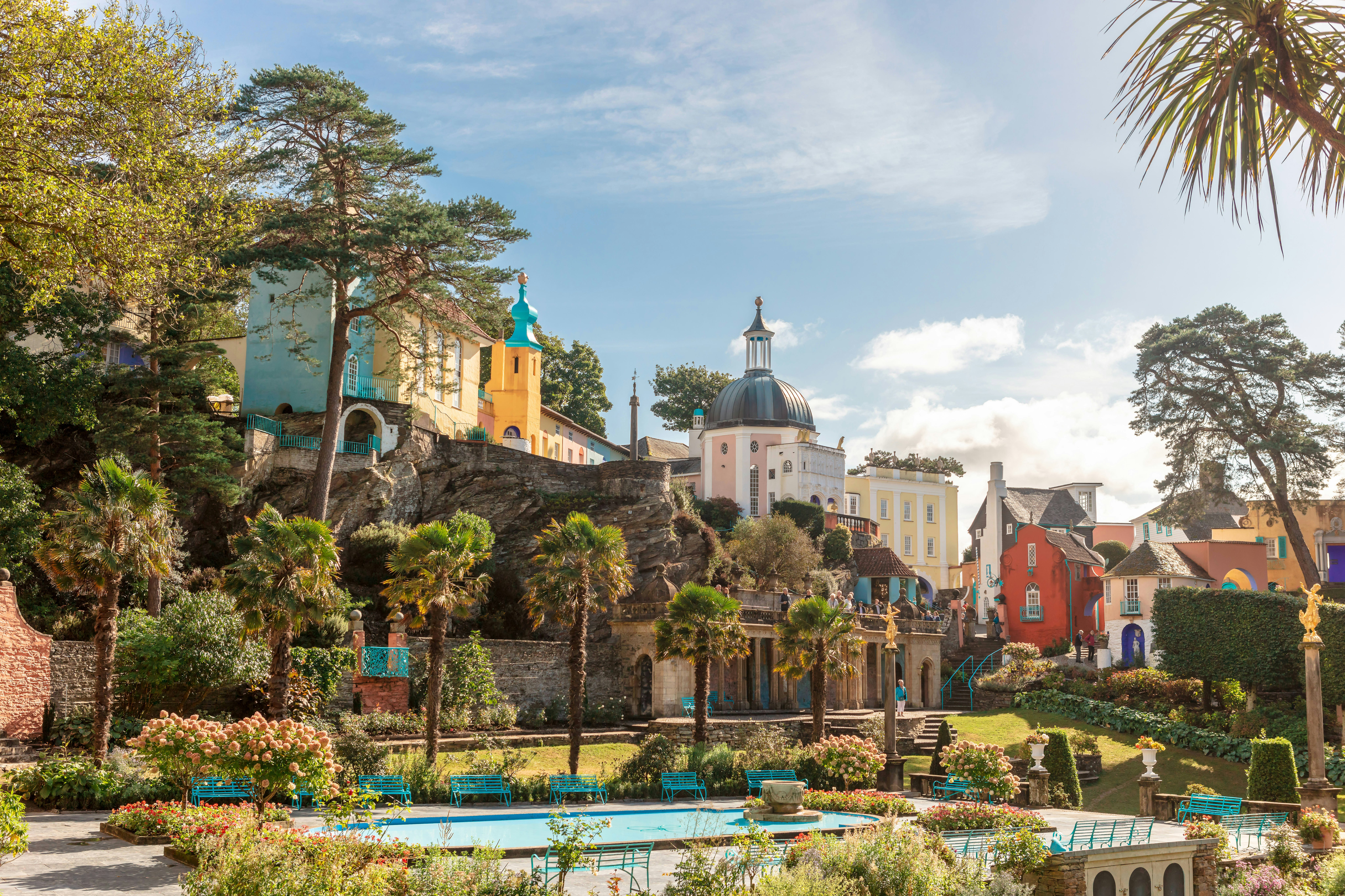 A park in Portmeirion; it has been planted with Mediterranean-style trees and plants, and it's surrounded by several elevated, brightly coloured buildings, giving the impression of an Italian hilltop town.