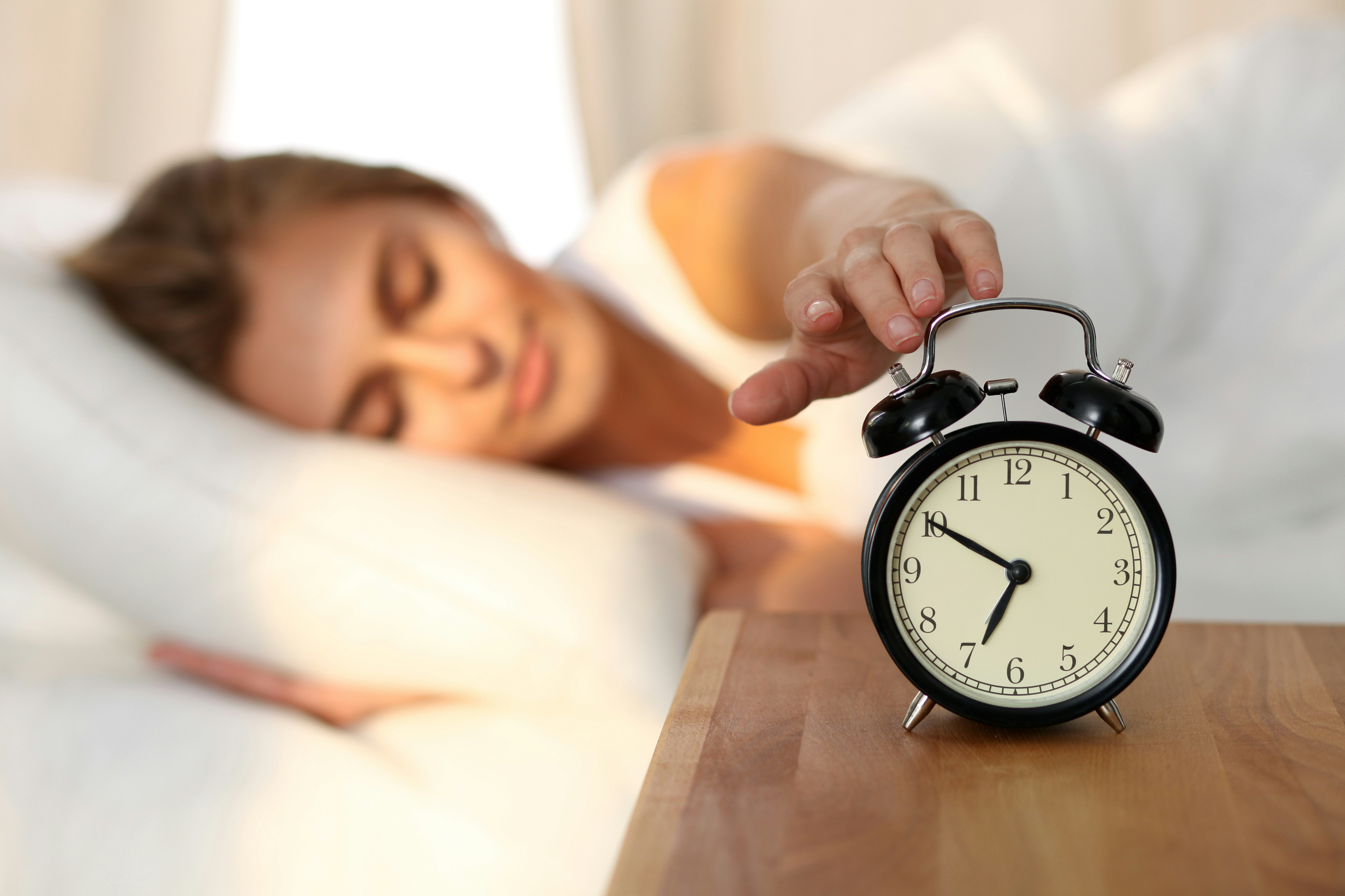 A woman in bed with her eyes closed reaches for her alarm clock 