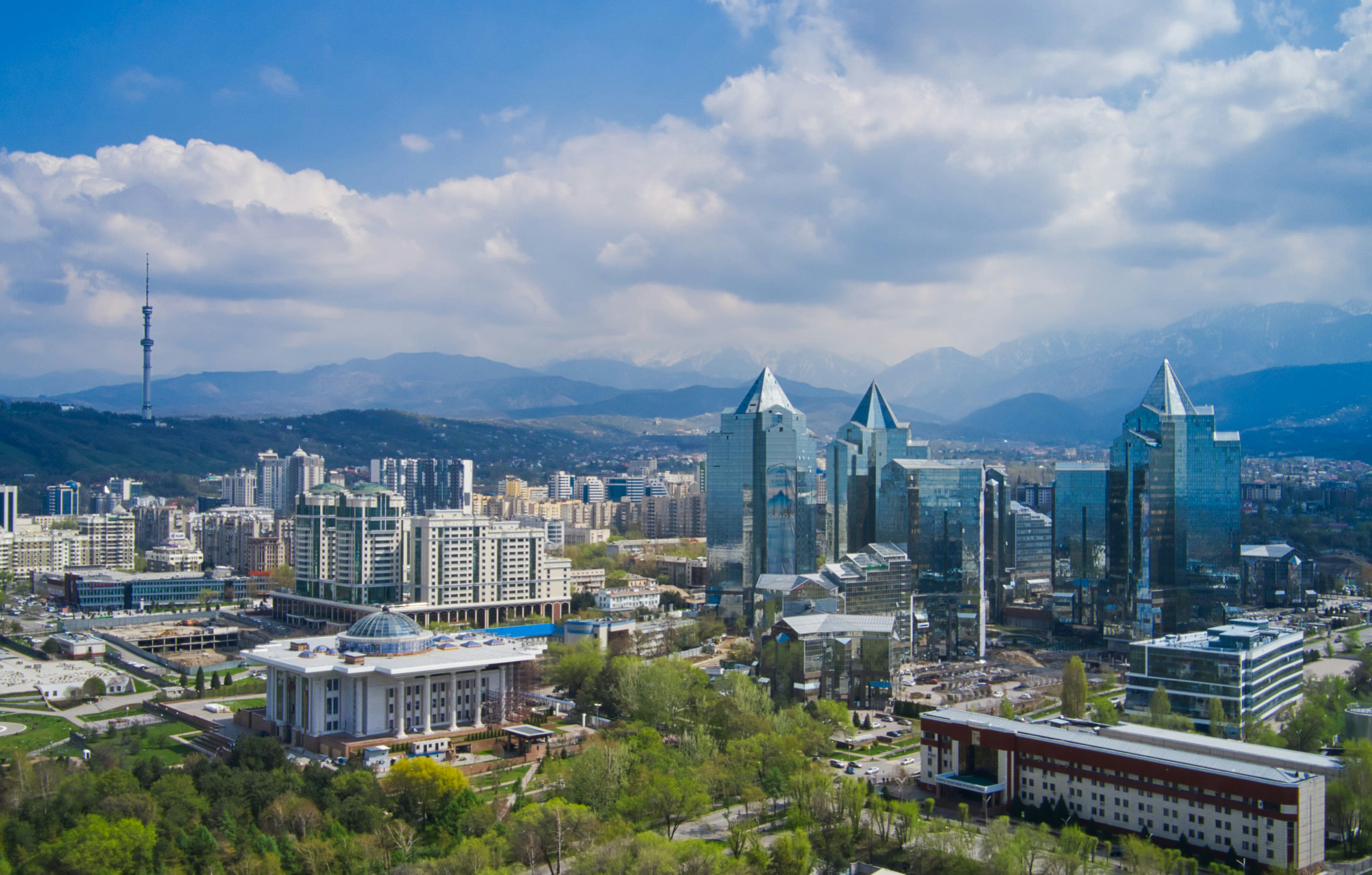 An aerial view of the Almaty skyline; showing modern skyscrapers, apartment blocks and green spaces backed by snow-capped mountains.