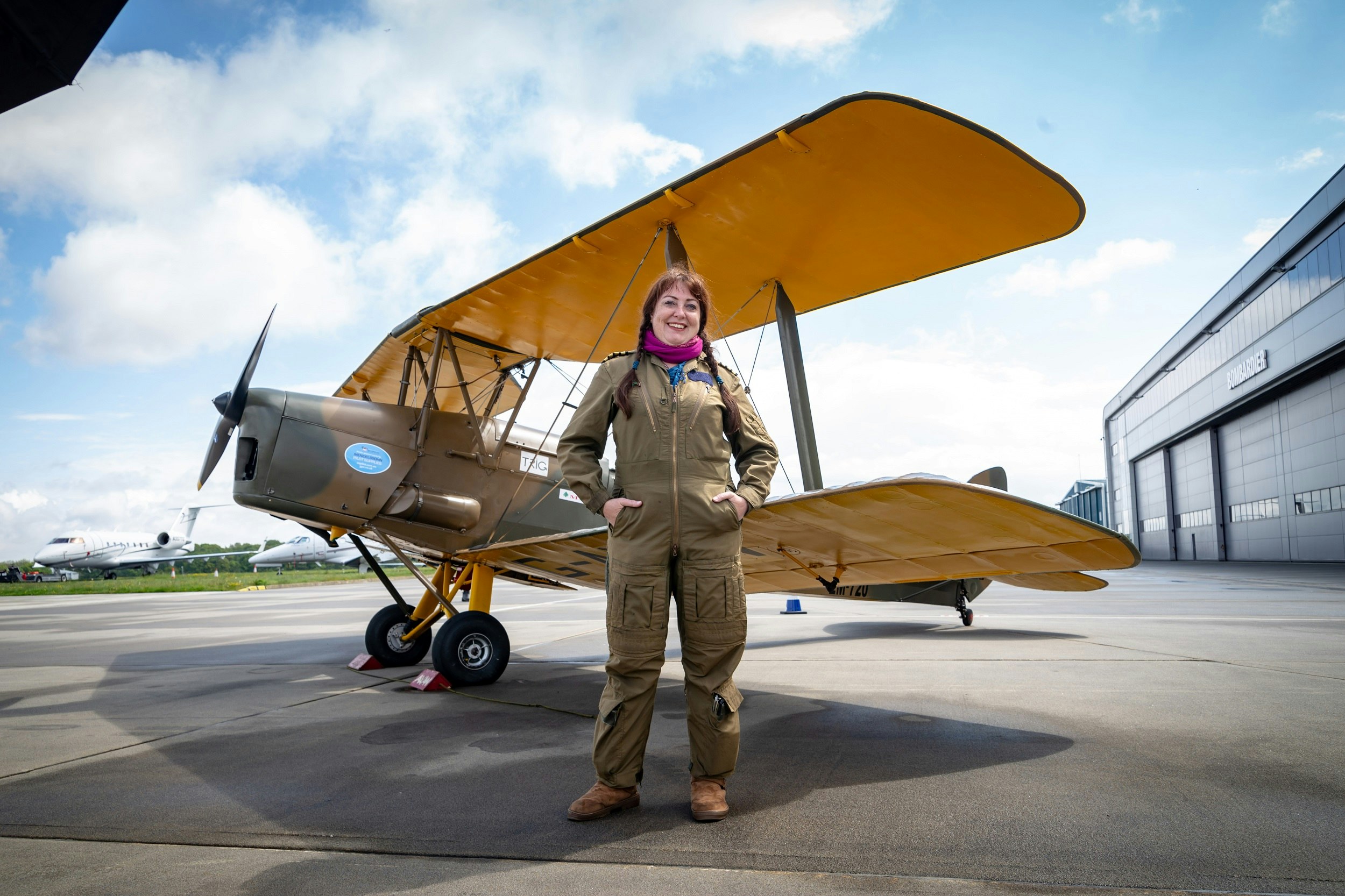 A female pilot in a flight suit stands beside her Tiger Moth plane at an airfield