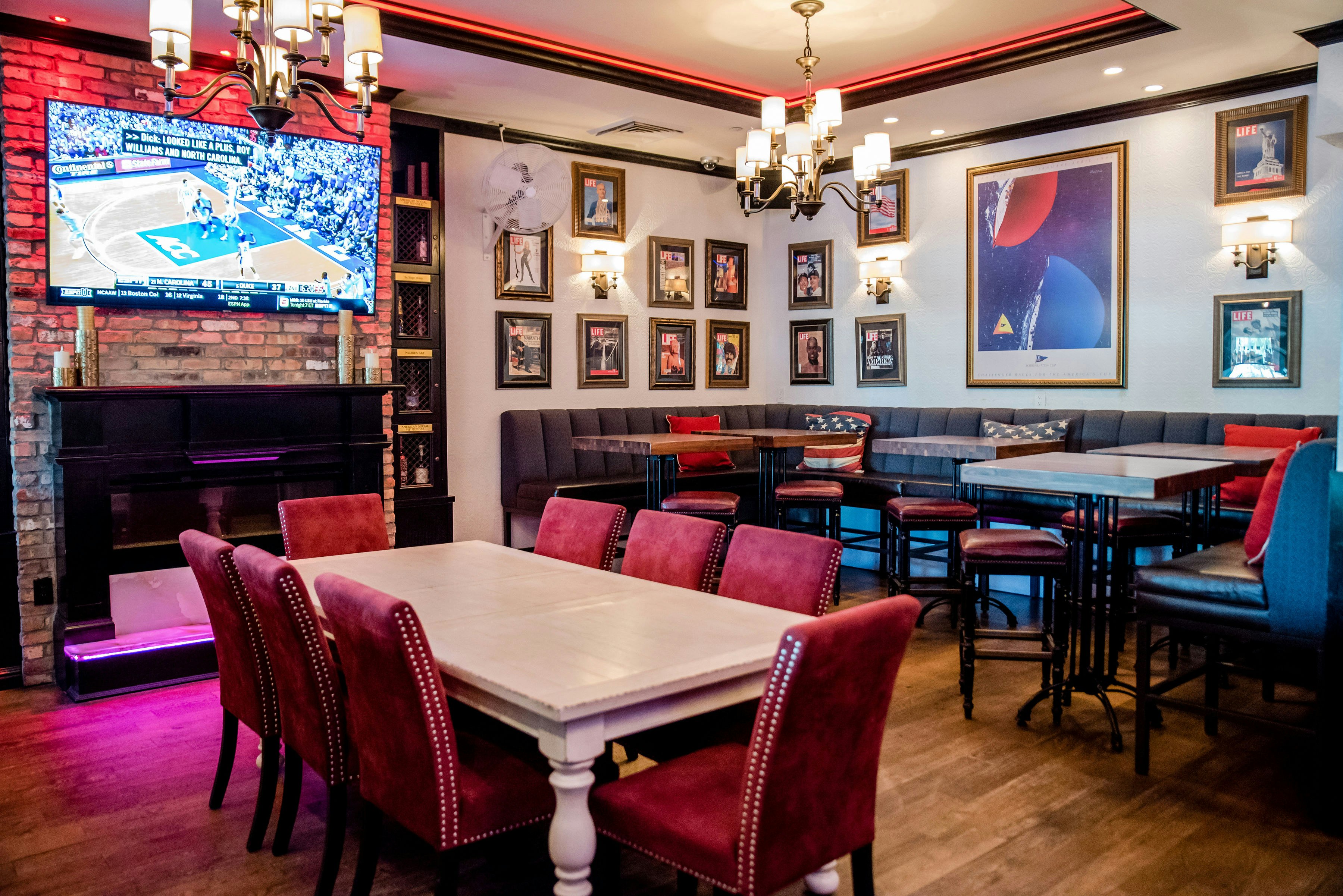 Interior of American Social Bar filled with plush chairs and wooden tables. There is a large TV on a brick wall above a faux fireplace.  