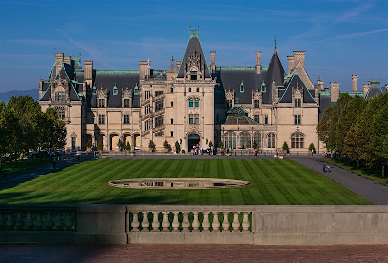 The Biltmore estate is a stately home in Asheville North Carolina. Its long yard frames a grey stone building with many gabled and peaked dark roofs; Best American architecture