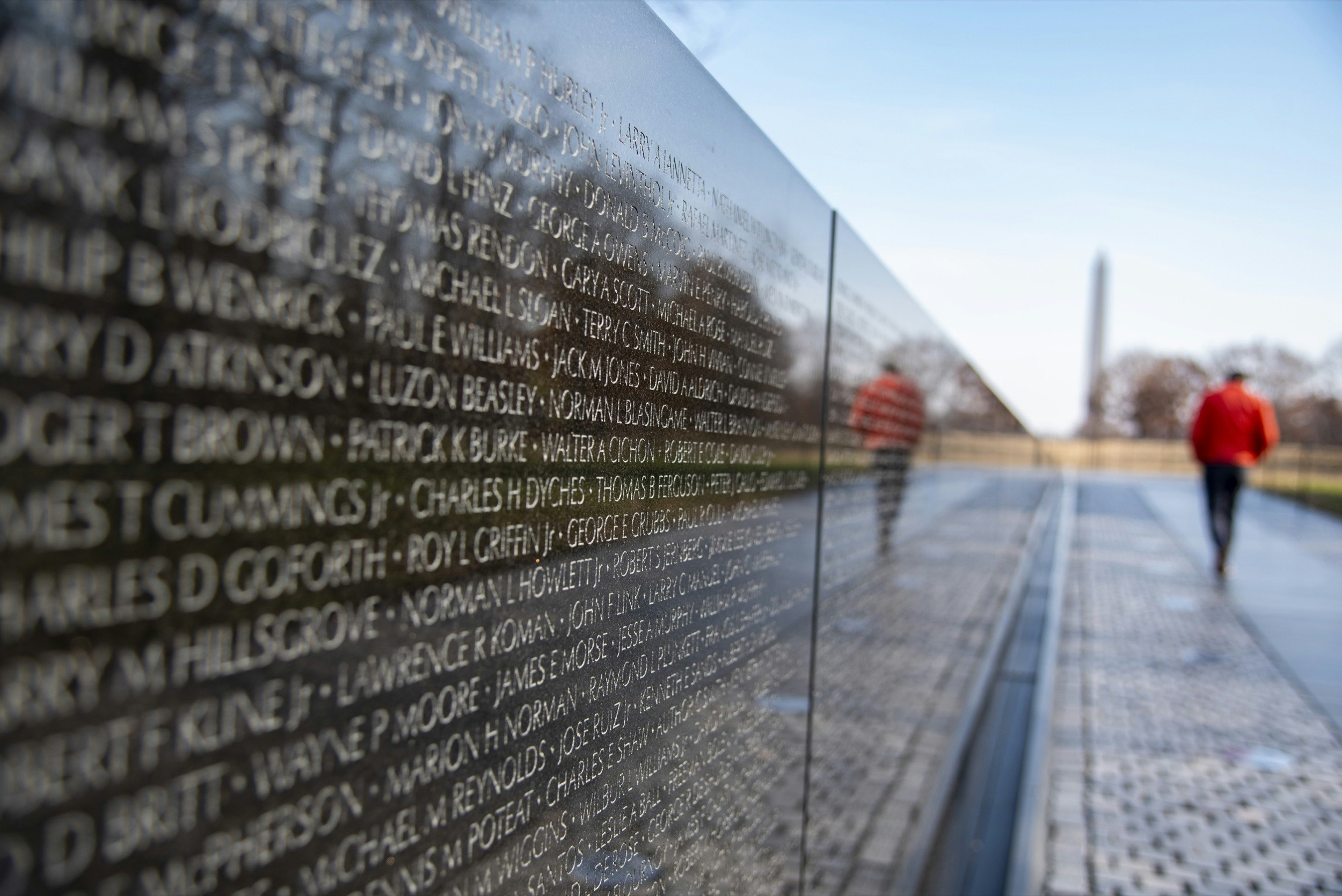 A man in a red coat is reflected in the black granite walls of the Vietnam Veterans Memorial as he walks toward the Washington Monument on a cold day. The names of deceased veterans are etched in the wall over his reflection; Best American architecture