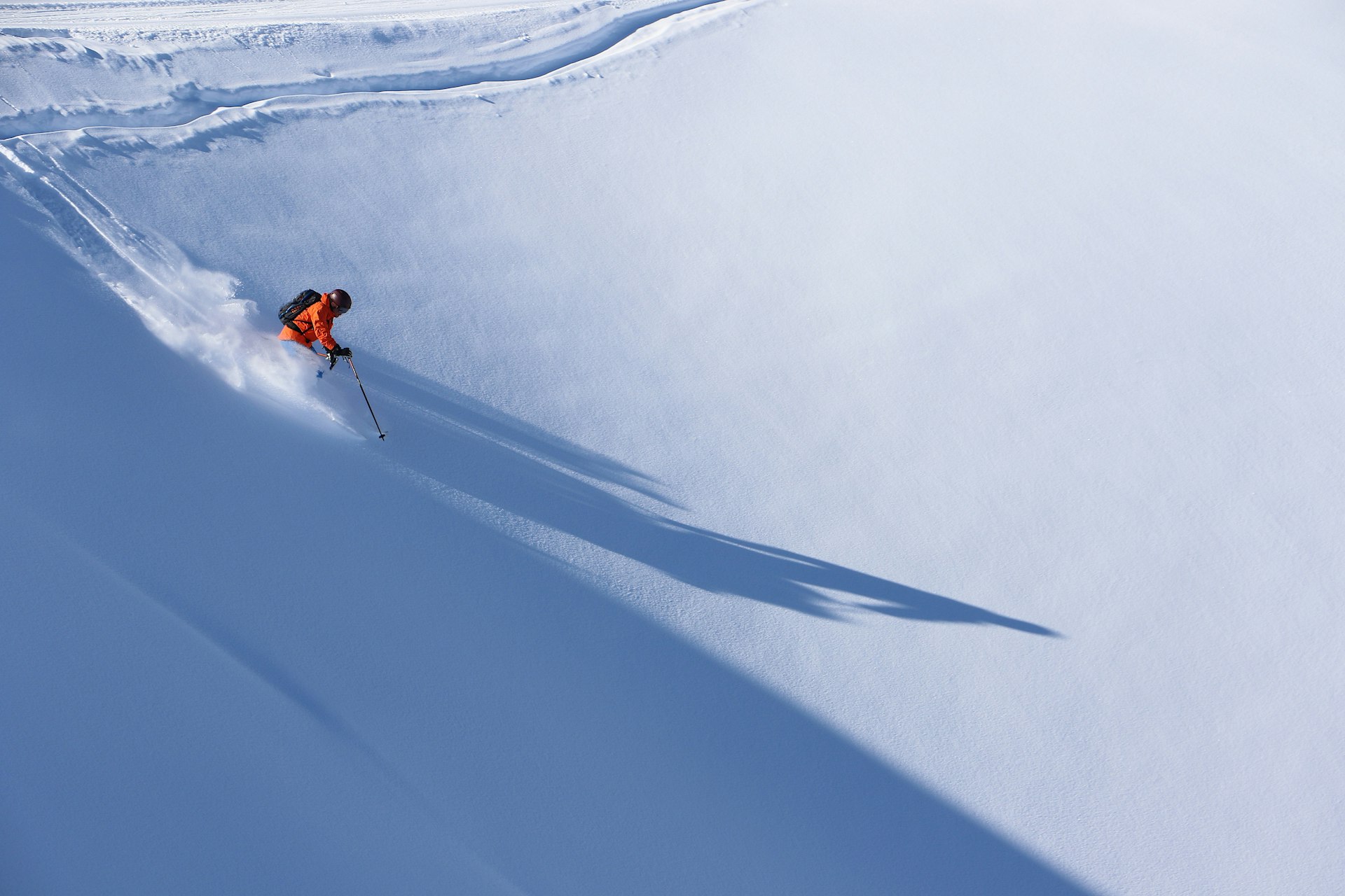 A single skier, dressed in a bright orange jacket, hurtles down a piste of perfect, untouched snow.