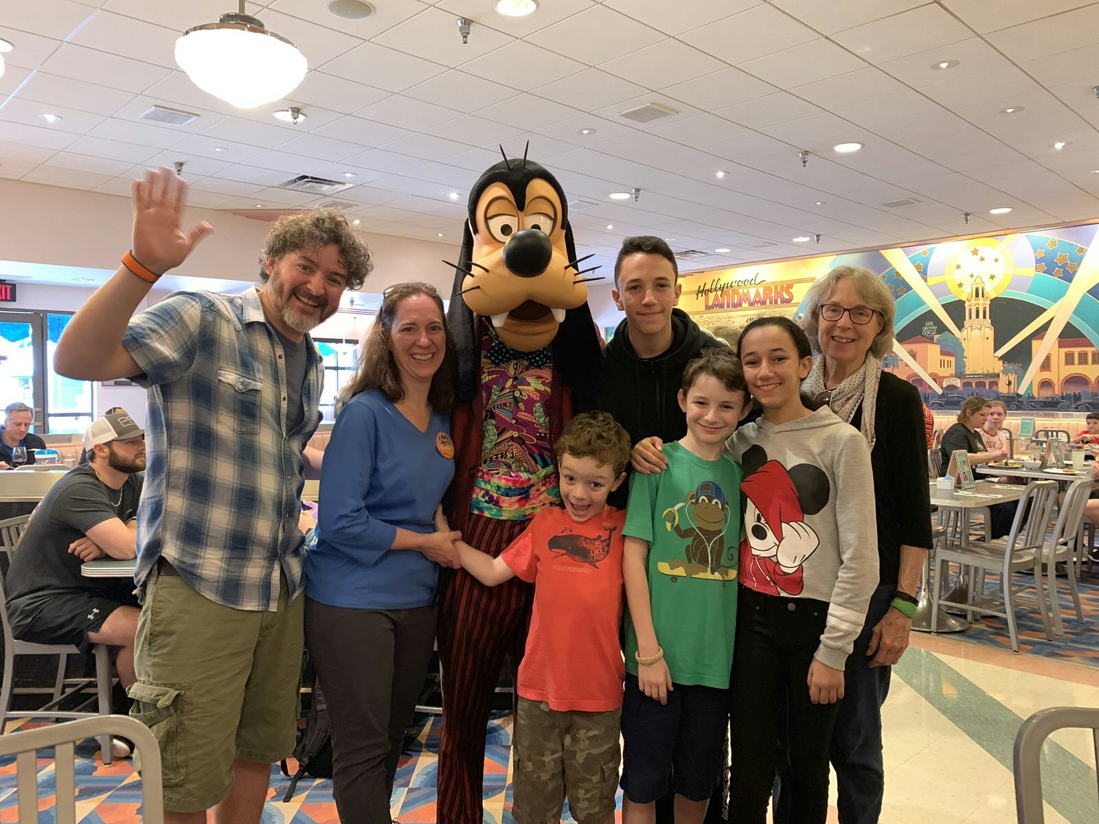 A family smile and wave at the camera gathered around someone in a Goofy costume at Disney World