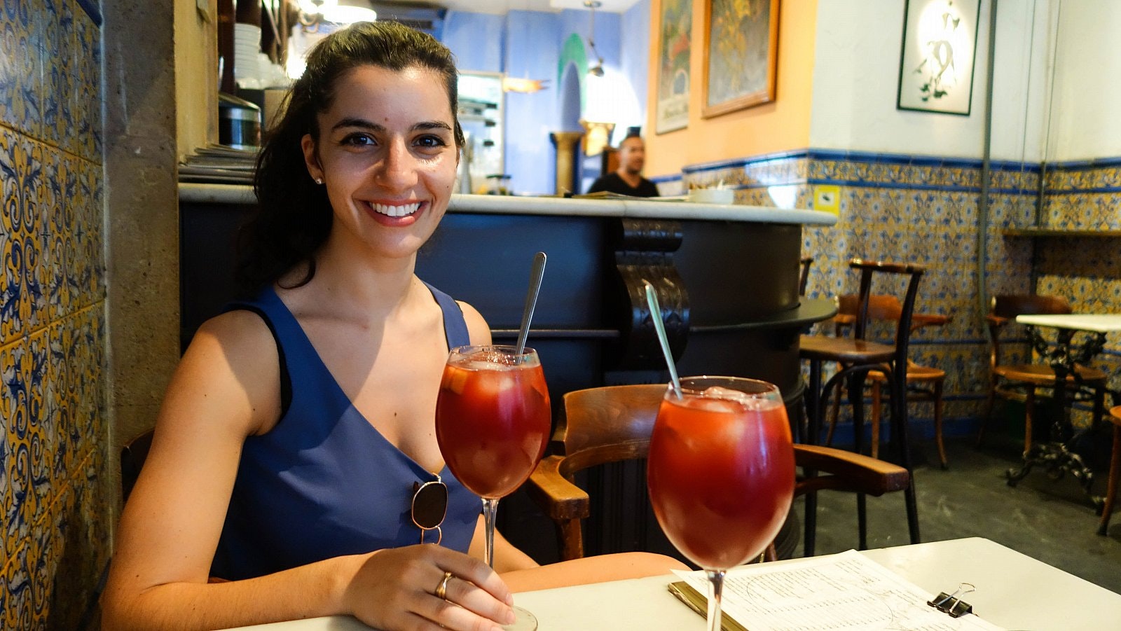 Caterina sits in a bar with tiled walls in a traditional yellow, white and blue geometric design; two full glasses of sangria are on the table in front of her.