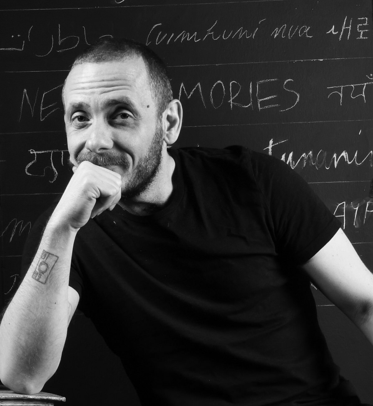 A black and white portrait of a man from the chest up, sitting in front of a blackboard, resting his head on his closed fist, looking into the camera