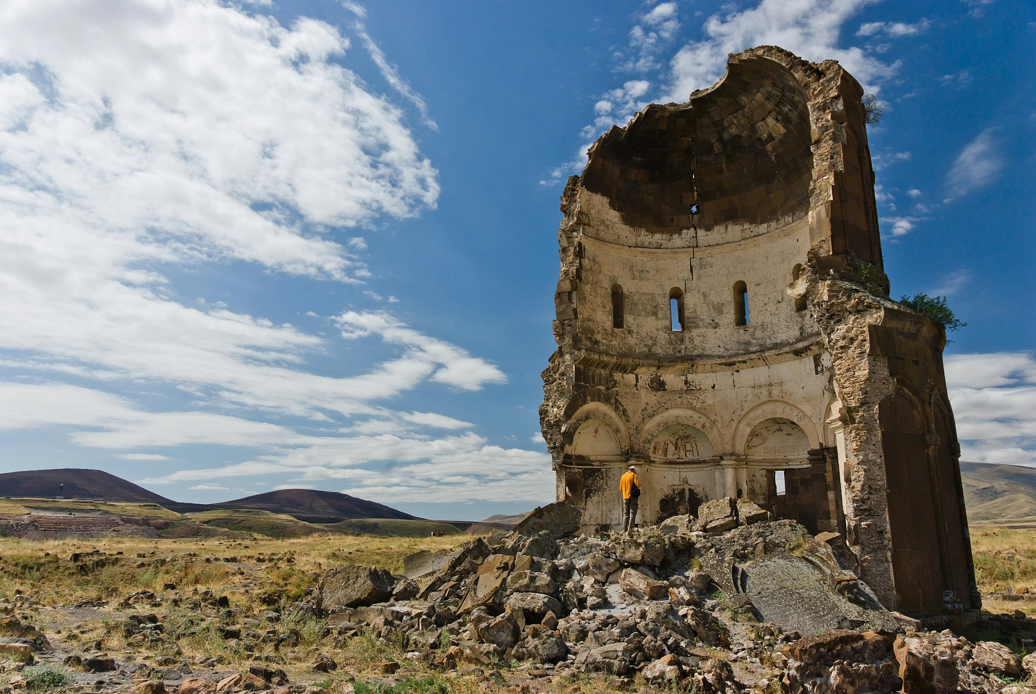 A person stands on a pile of rubble looking inside the remains of a half-destroyed domed church, with a grassy plain and hills in the background.