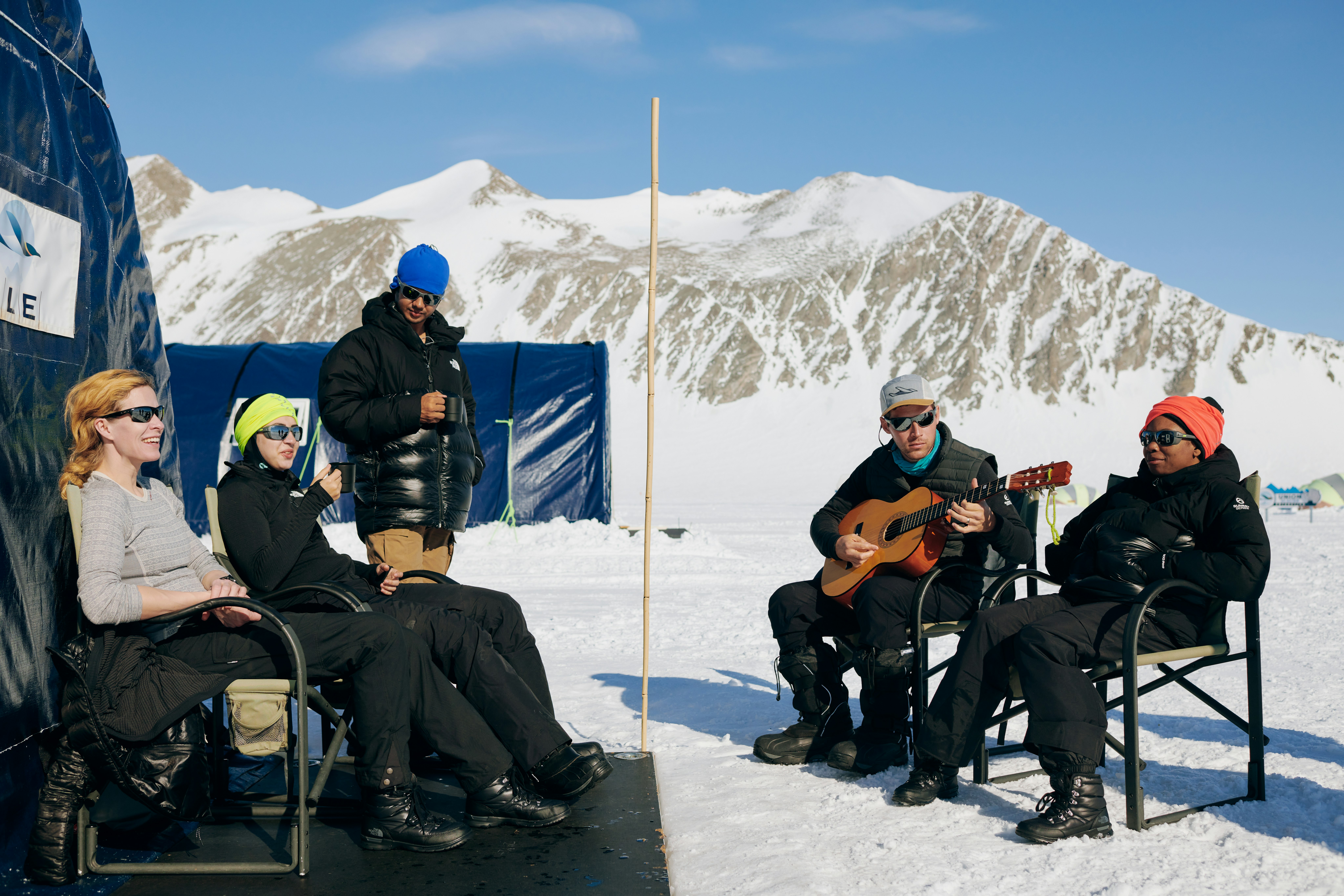 A group of volunteers sit on chairs and play guitar outside the tents of their Antarctica base camp