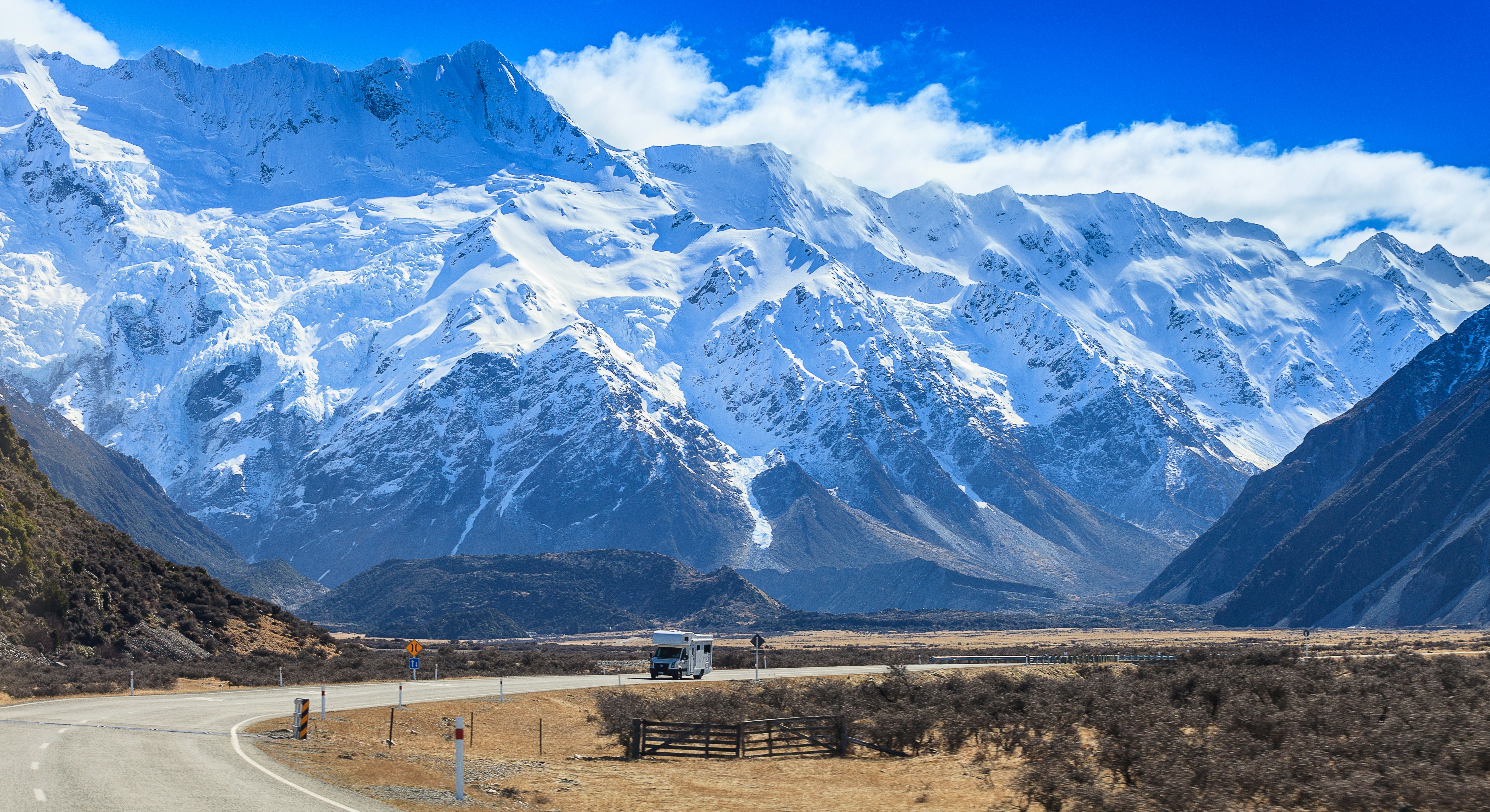 A massive snow-capped mountain range towers over a road. A camper van is the only vehicle on the road