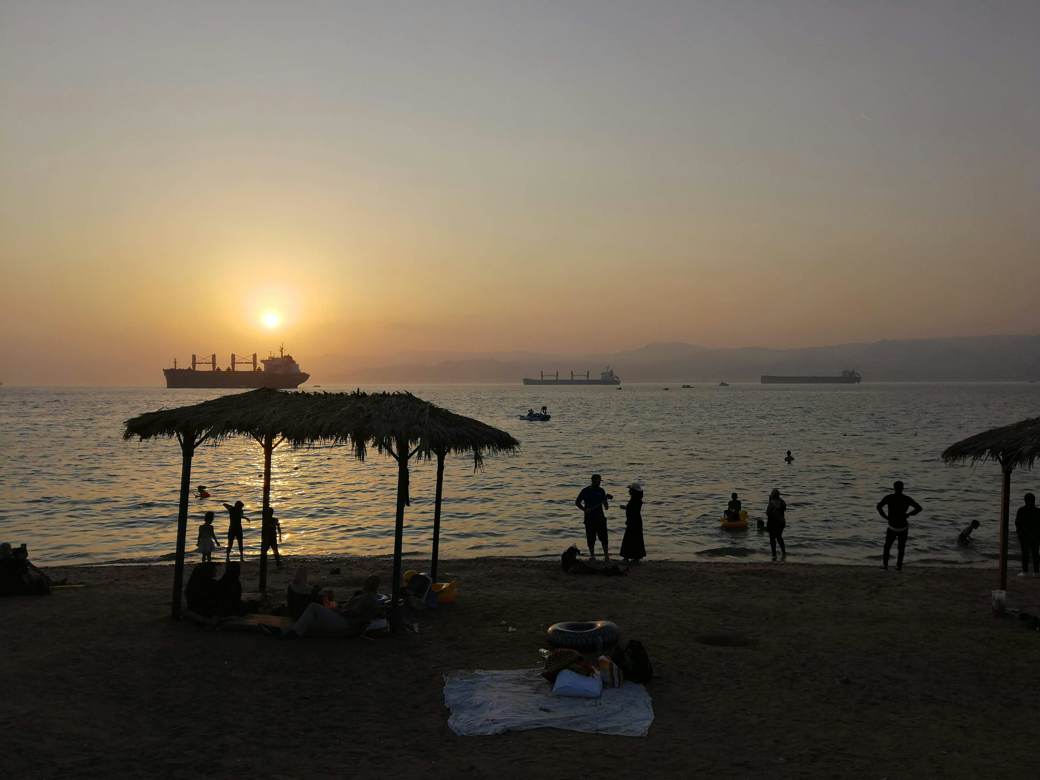 A beach at sunset. In silhouette are children playing in the water, a palm tree shelter and adults standing on the shore. A ship is on the water.