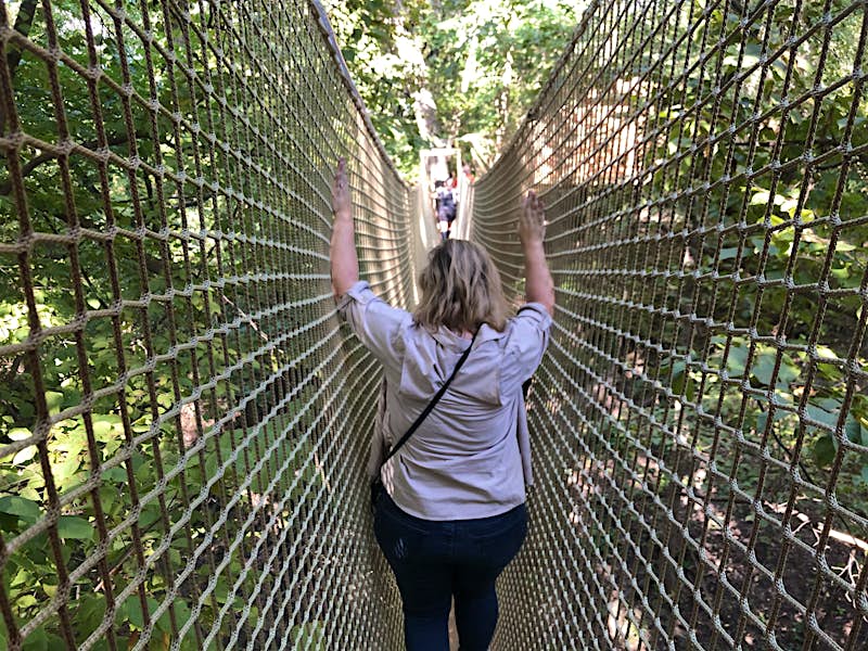 A woman walks along a narrow rope bridge that has high netting on either side; her up-stretched hands are still a few feet below the top of the netting.