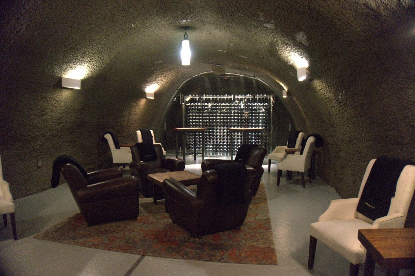 The dark, masculine interior of Archery Summit Tasting Room is a curved, arch-shaped space with dark walls and white lights highlighting a red and brown rug, dark leather arm chairs, and white contemporary chairs