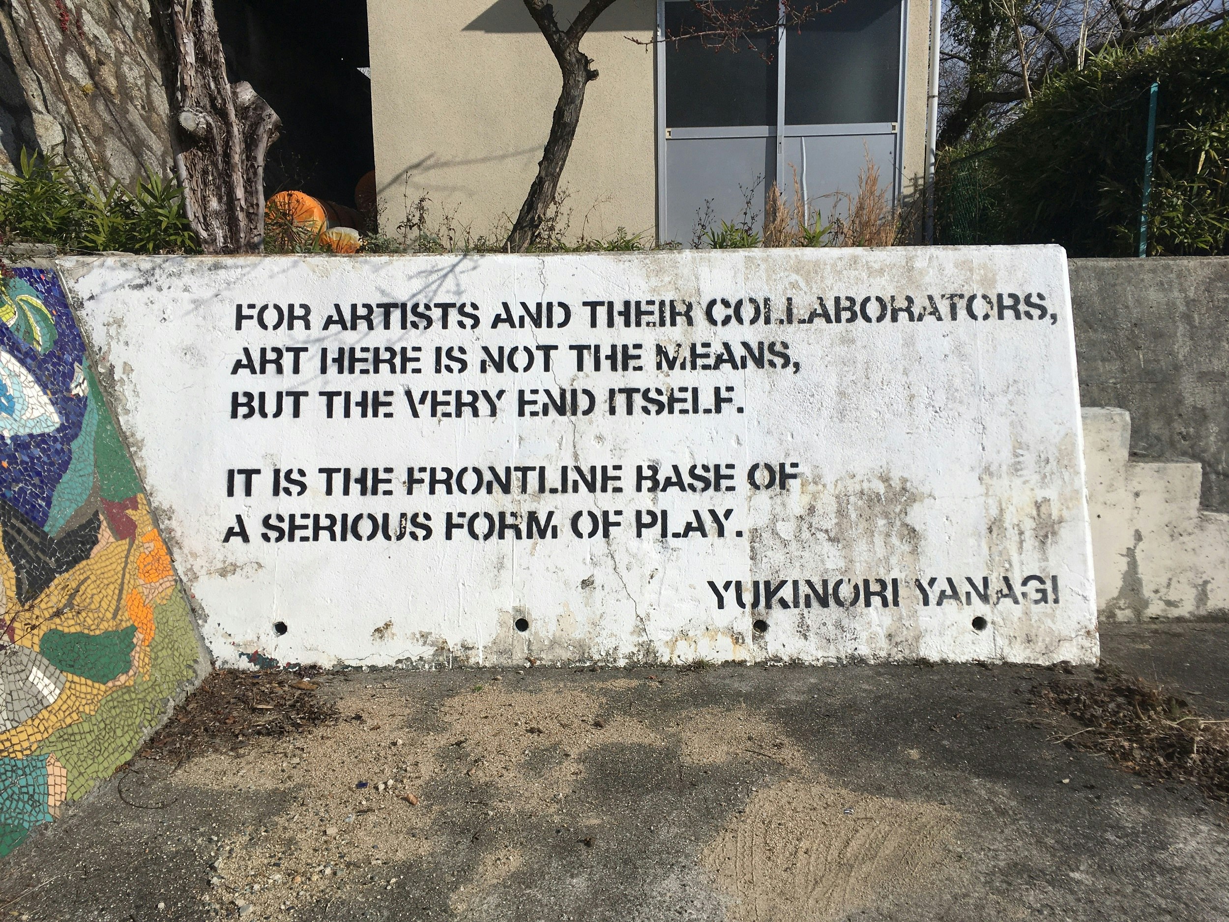 Words painted in black on a white wall, which read: "For artists and their collaborators, art here is not the means, but the very end itself. It is the frontline base for a very serious form of play. Yukinori Yanagi".