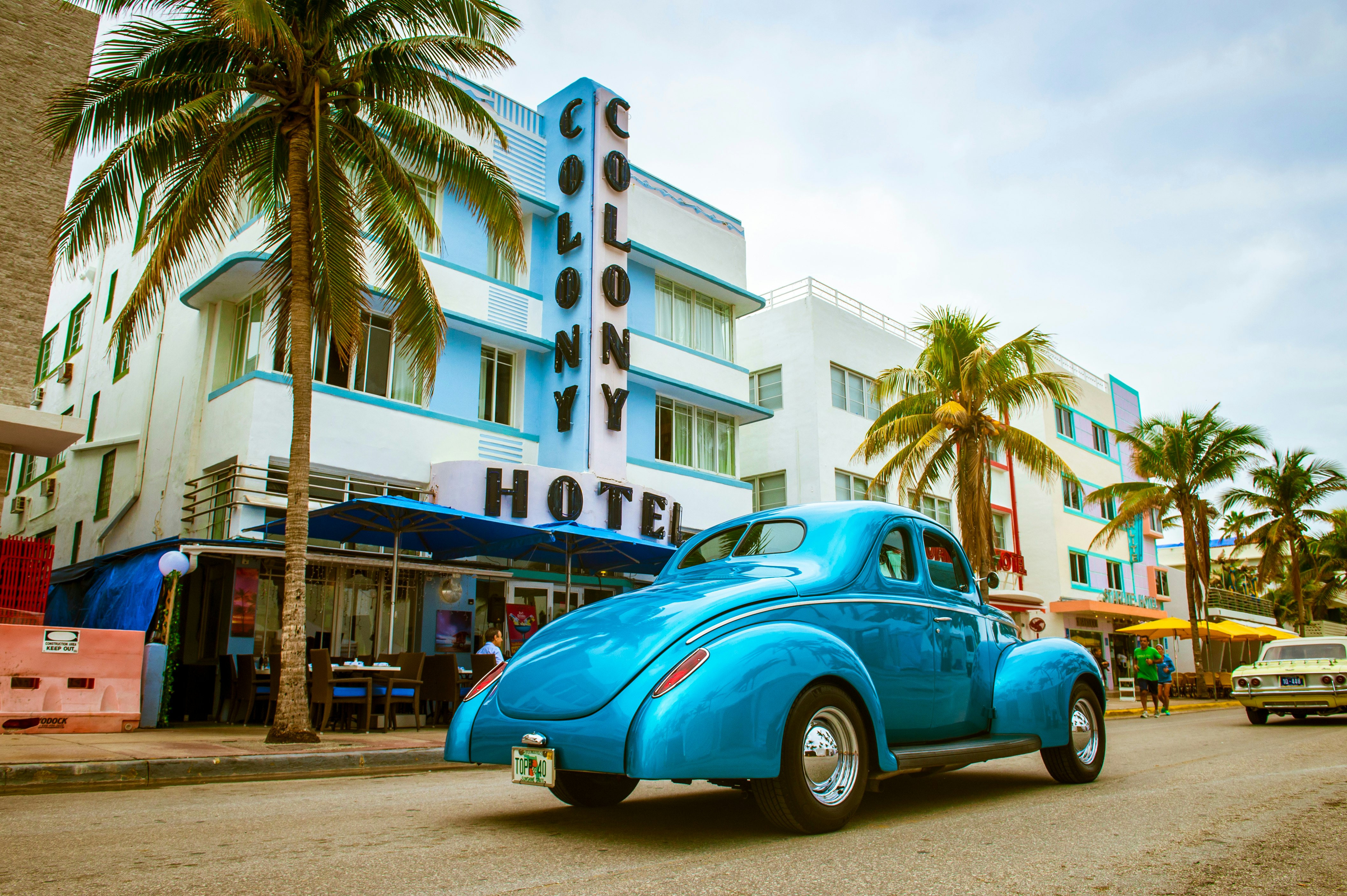 A light blue vintage car is parked outside of the Art Deco-inspired Colony Hotel in Miami. The hotel is flanked by palm trees. 