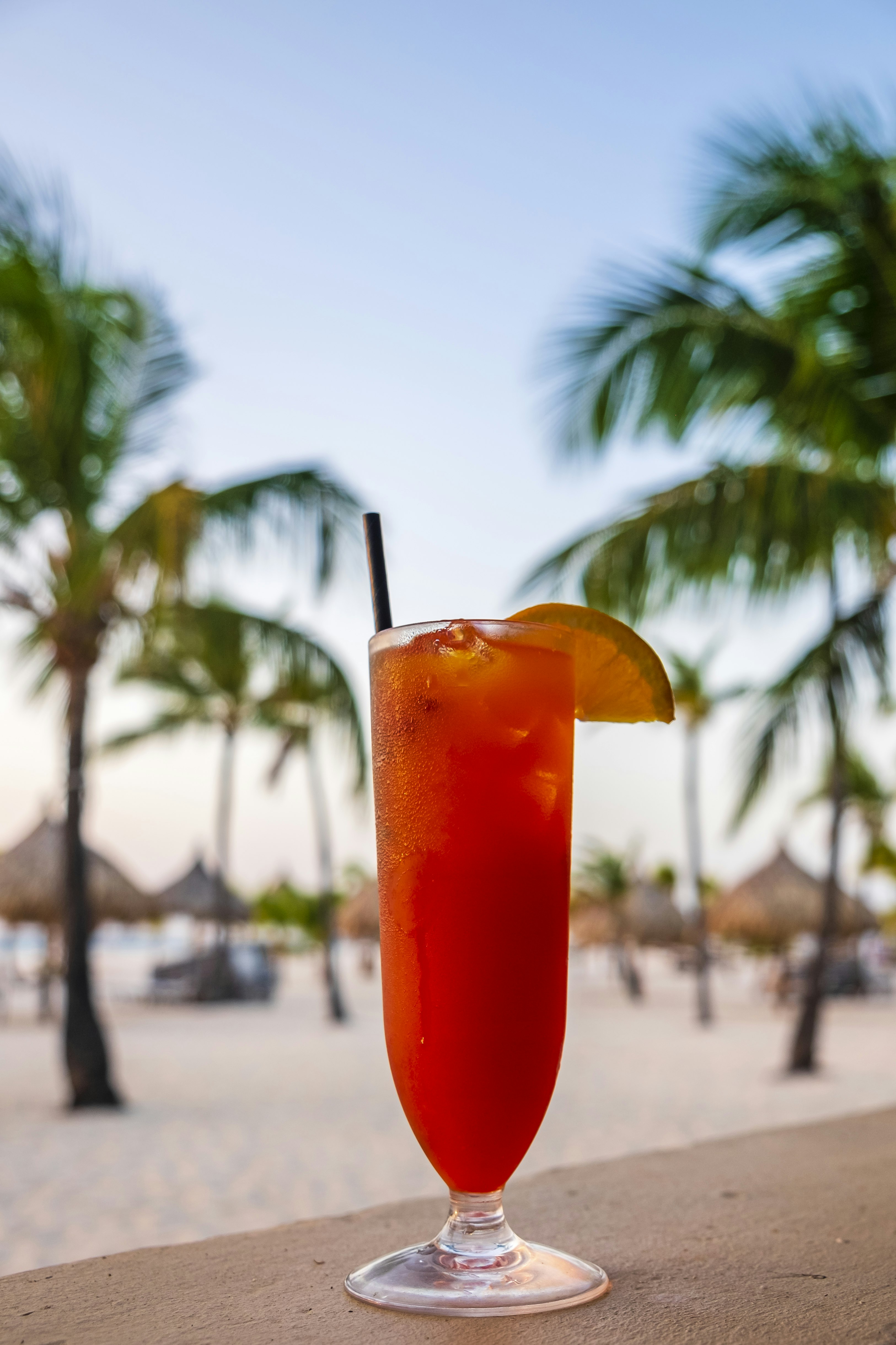 A tall glass of an Aruba Ariba on ice, garnished with a lime wedge and a black straw rests on a cement ledge. In the background is sand, small huts and palm trees.