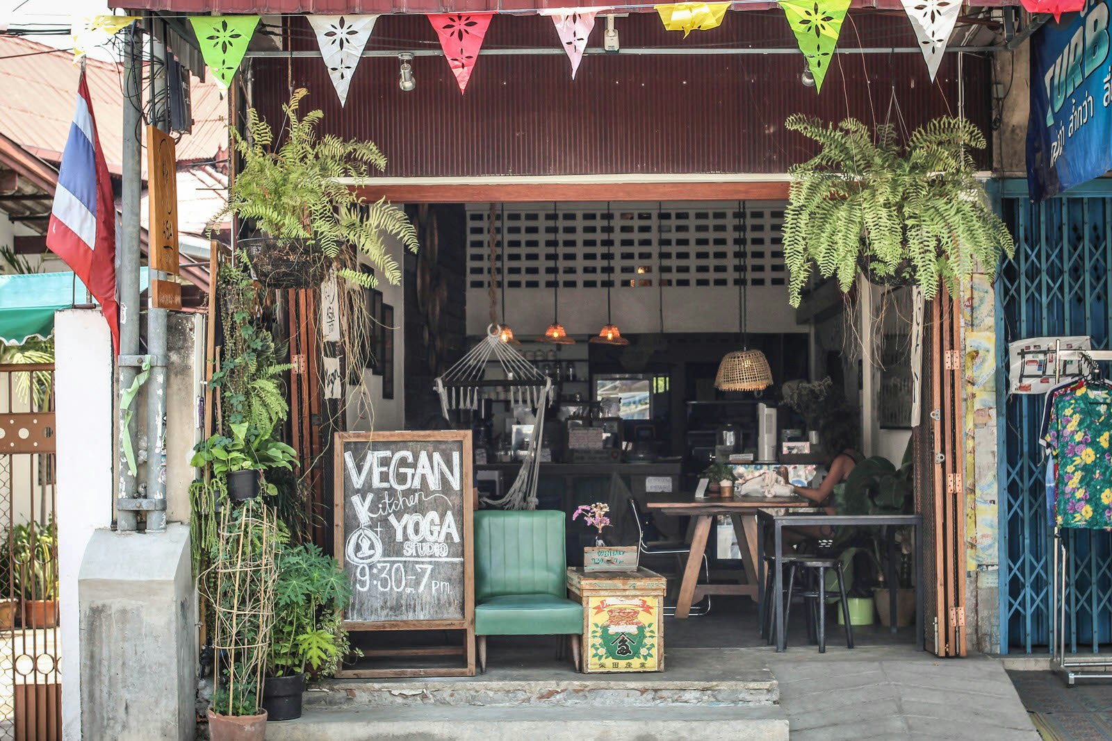 The exterior of Asa cafe in Chinag Mai. There is colourful bunting and hanging plants hung in front of the cafe and a blackboard reading 'VEGAN Kitchen + YOGA STUDIO' with class times underneath. There's comfy seating and a cafe counter visible inside. 