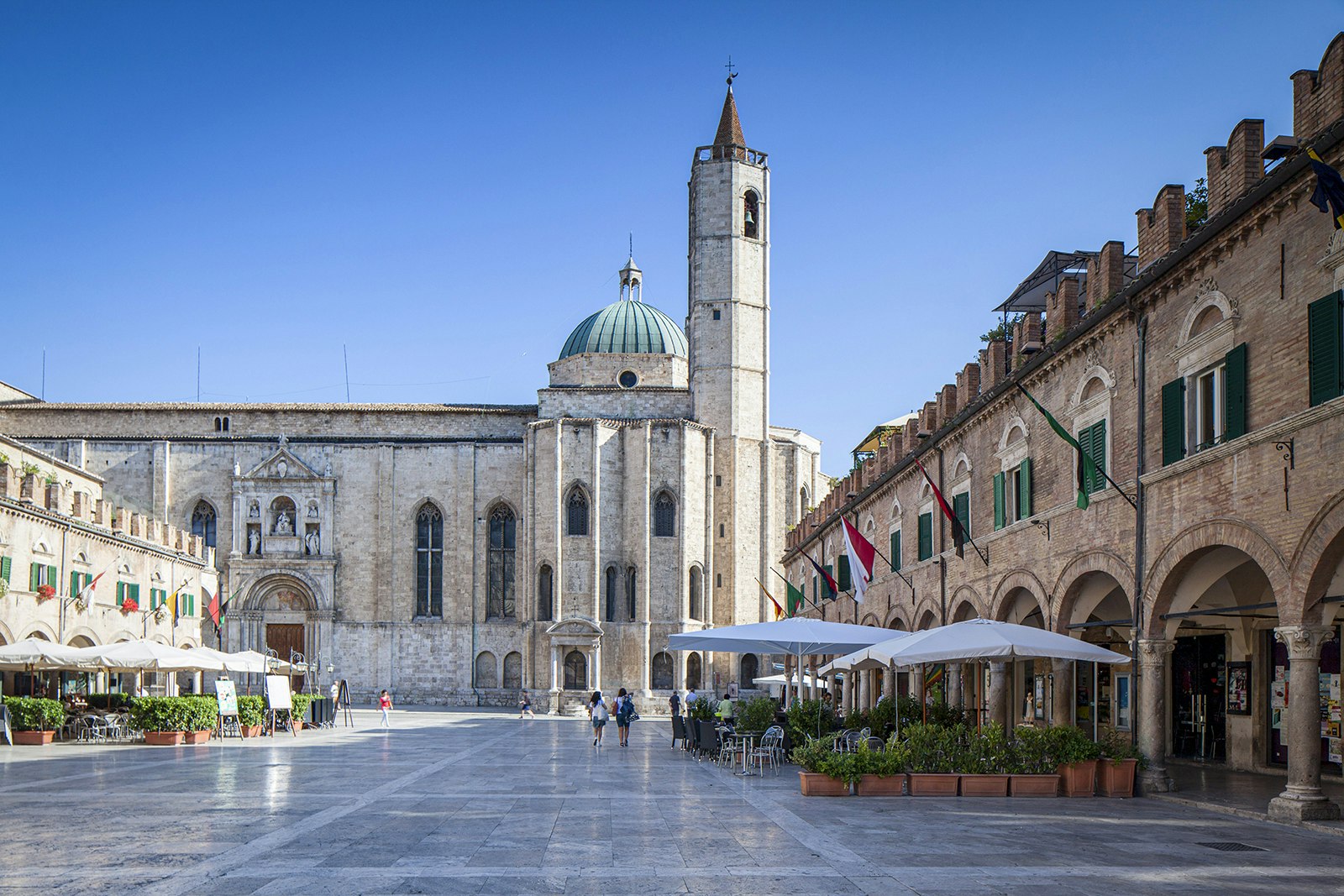 People walk through a stone-tiled piazza backed by a cathedral in Ascoli Piceno. The piazza is lined by historic buildings and bar terraces.
