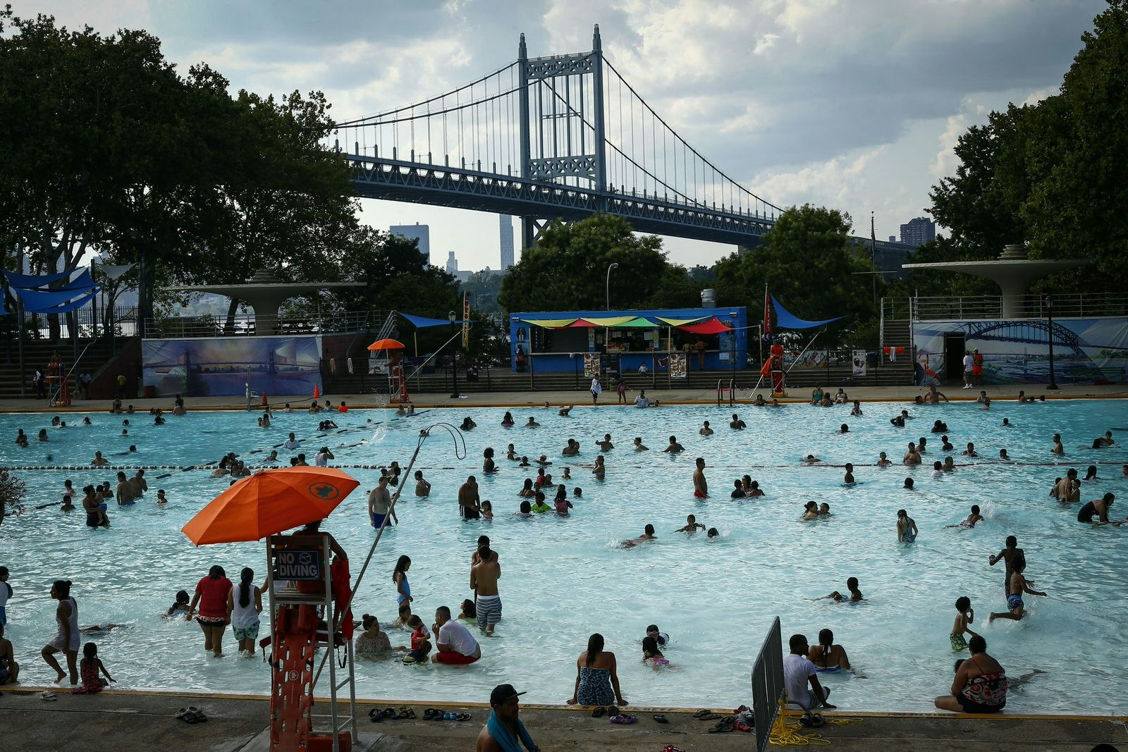 People enjoy a warm day at the Astoria Park Pool in New York with the RFK bridge visible in the background.
