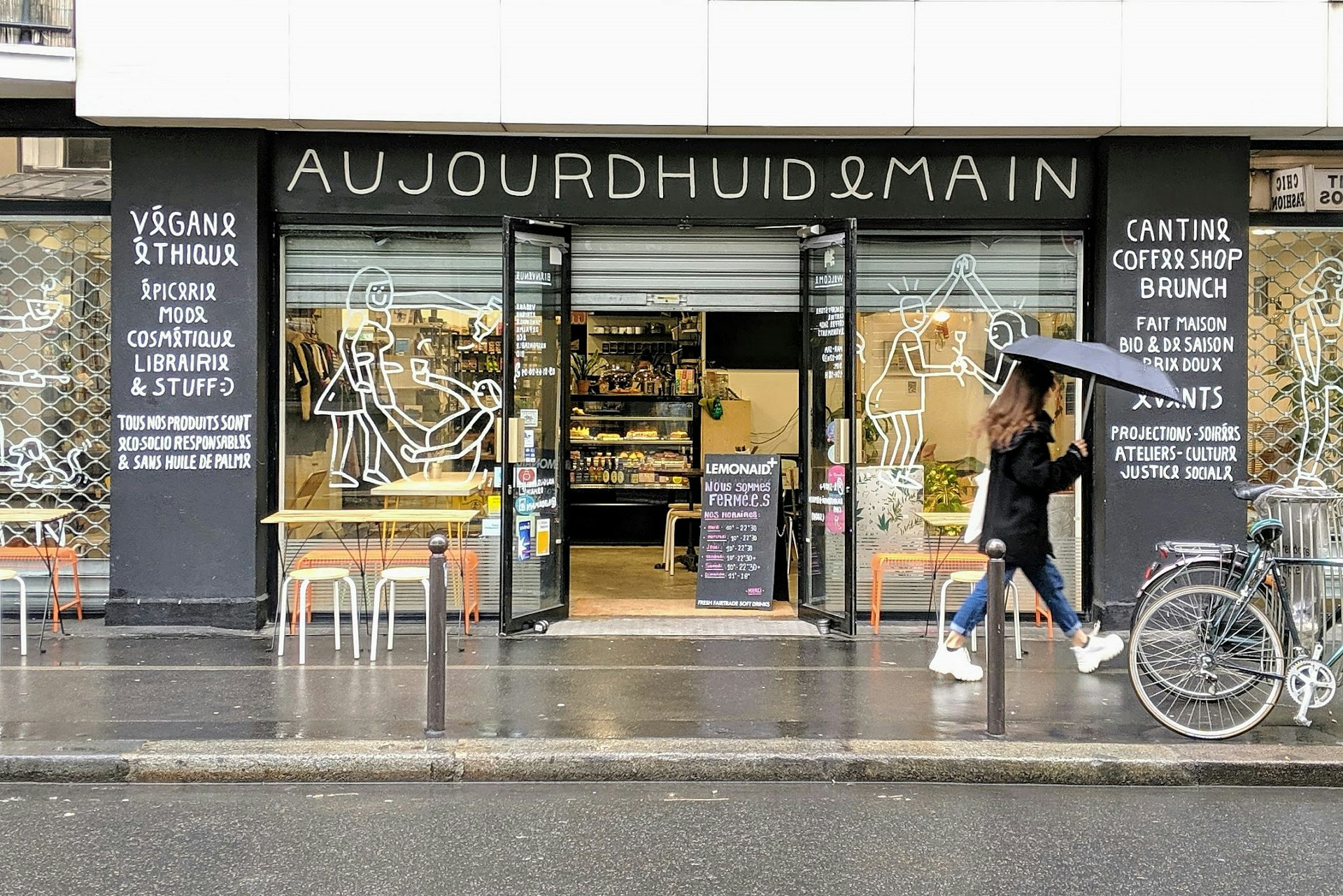 A lady with an umbrella walks past the Audjourd'hui Demain storefront; the walls are black with white signage and the large windows have white line drawings. 