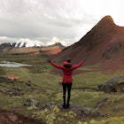 A woman in a red and black jacket extends her arms as she looks over a vast mountain landscape. 