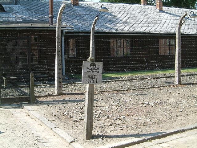 A sign with a skull and crossbones stands between two sets of wire fencing. The text says "Halt! Stoj!" 