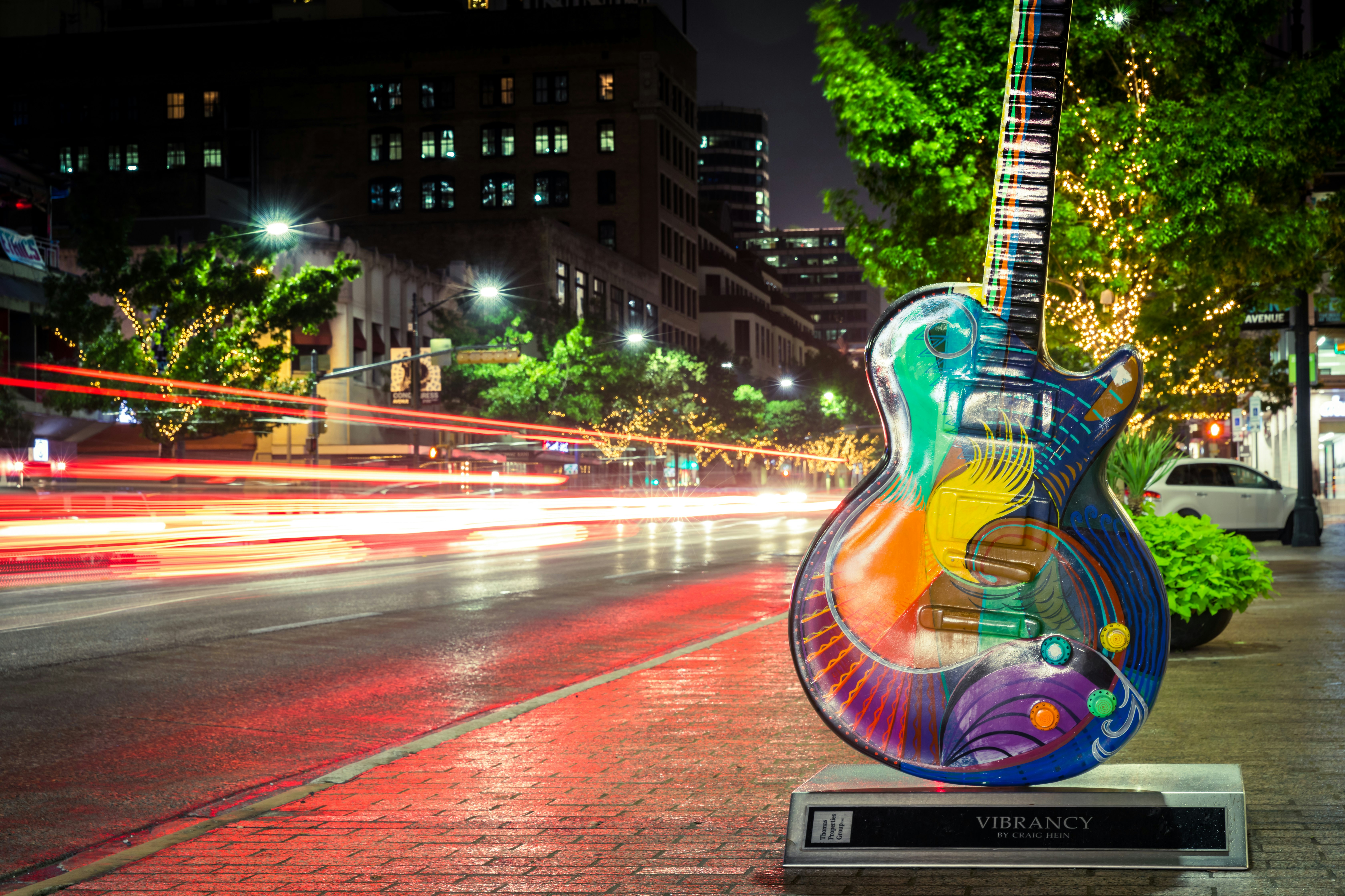 A large multi-colored guitar statue is erected next to a city street in downtown Austin. A blurry of cars zip by; Austin vs Norman