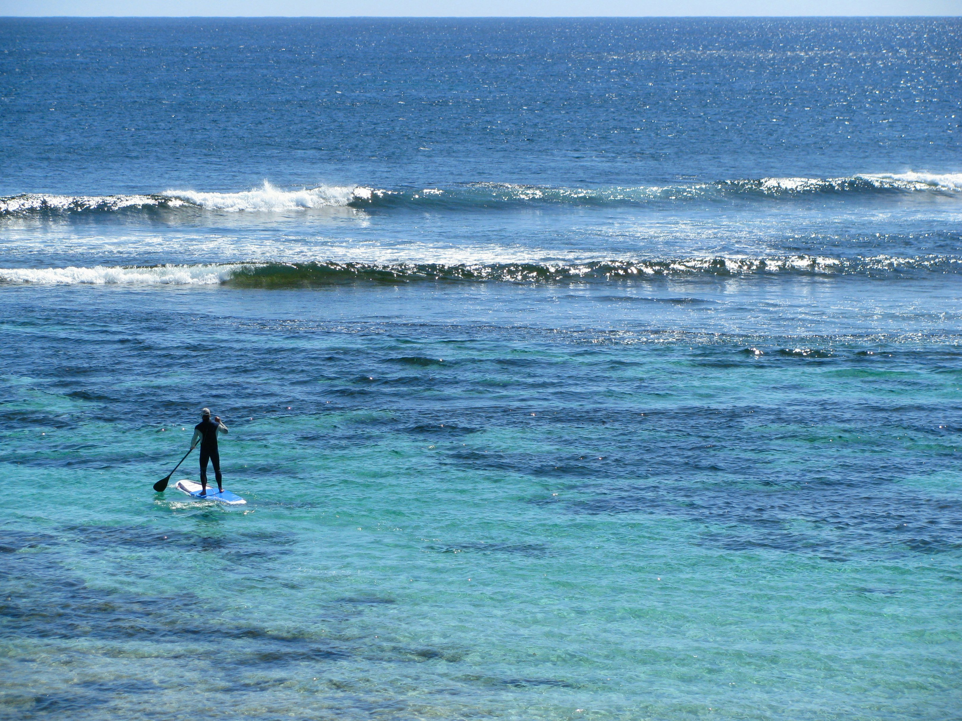 View of the back of a man in the distance, standing on a paddle board, paddling out alone into the ocean towards the waves; places for stand up paddle boarding