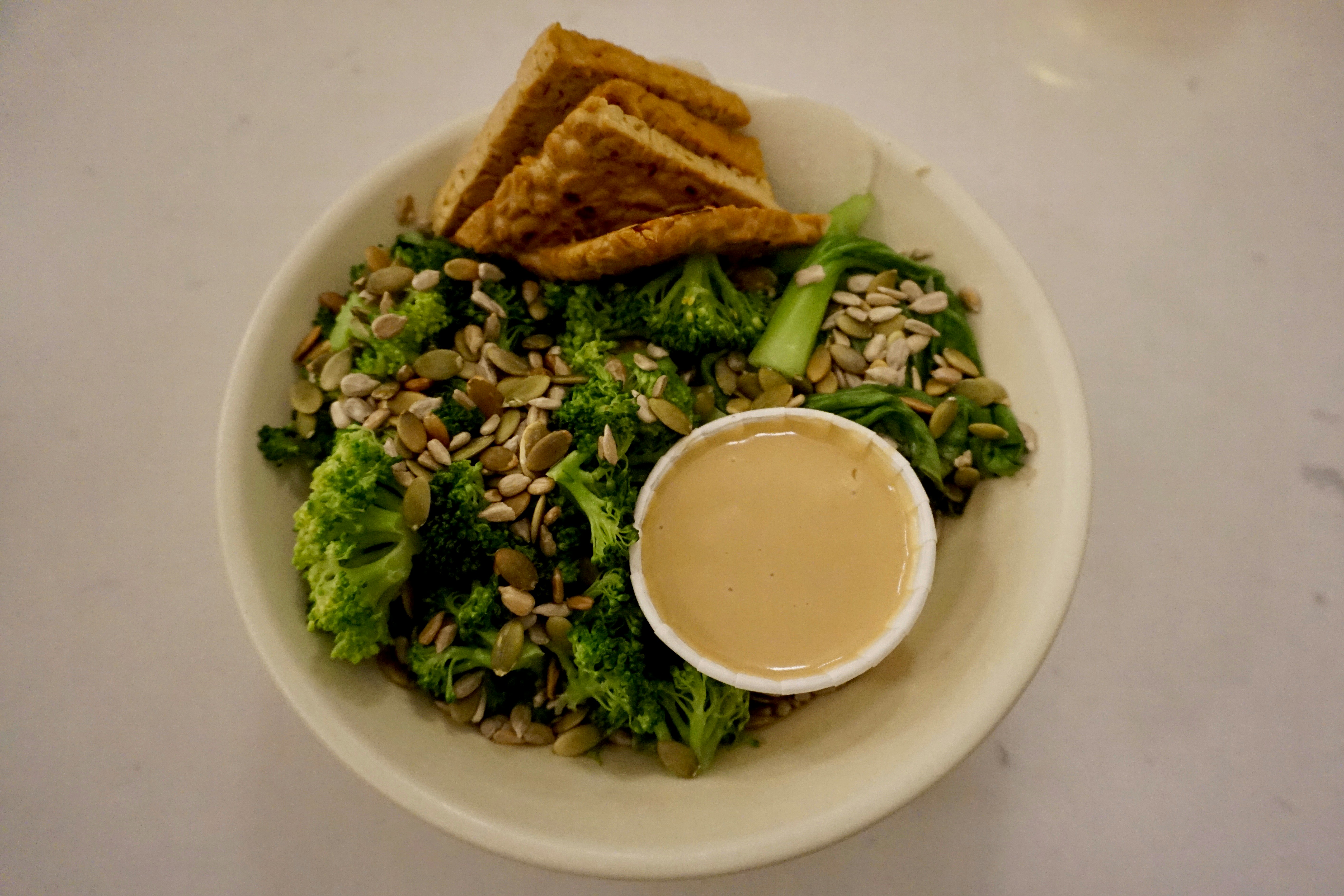 A white bowl, filled with broccoli, nuts, seeds, flatbread and a small container of satay sauce.