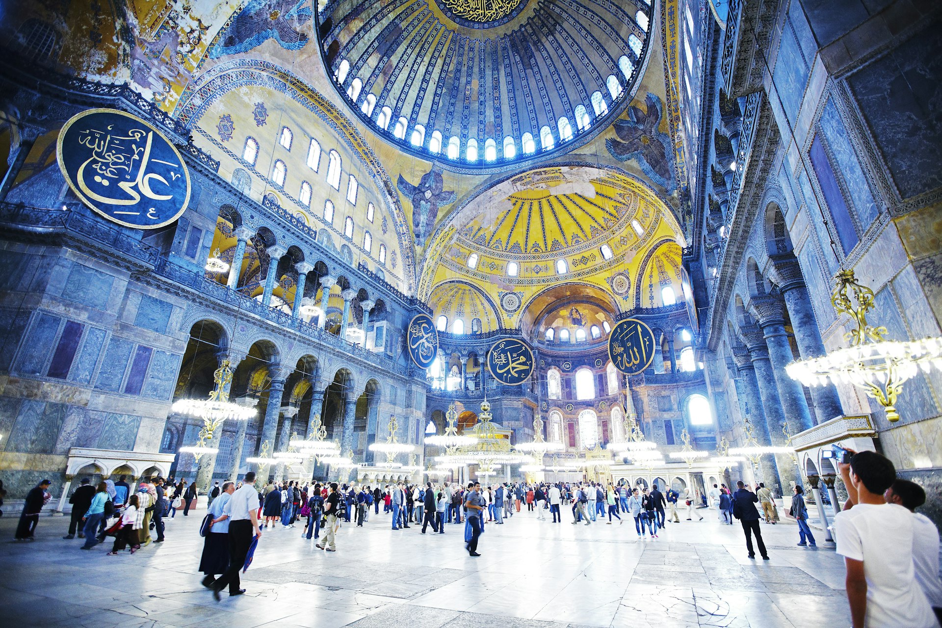 The light blue interior of Aya Sofya is highlighted with golden accents throughout the structure. There are large blue and gold signs attached to pillars around the building.