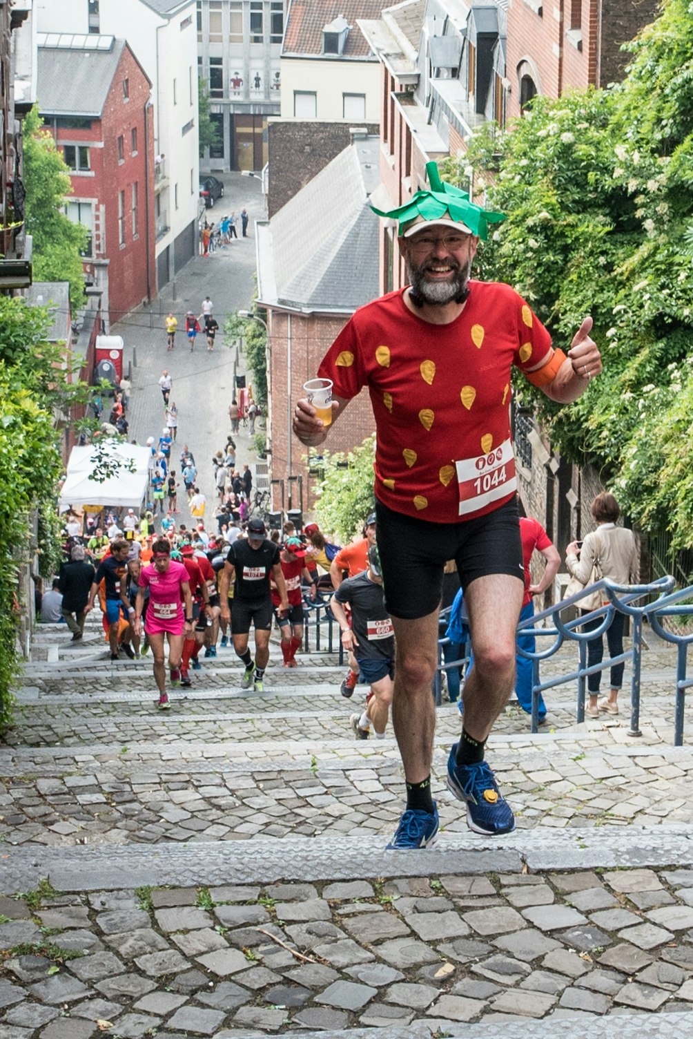 Many runners are climbing a steep set of stairs in a city. In the foreground, a man dressed as a strawberry is smiling at the camera, holding a tumbler of beer in one hand and giving the thumbs-up sign with the other.