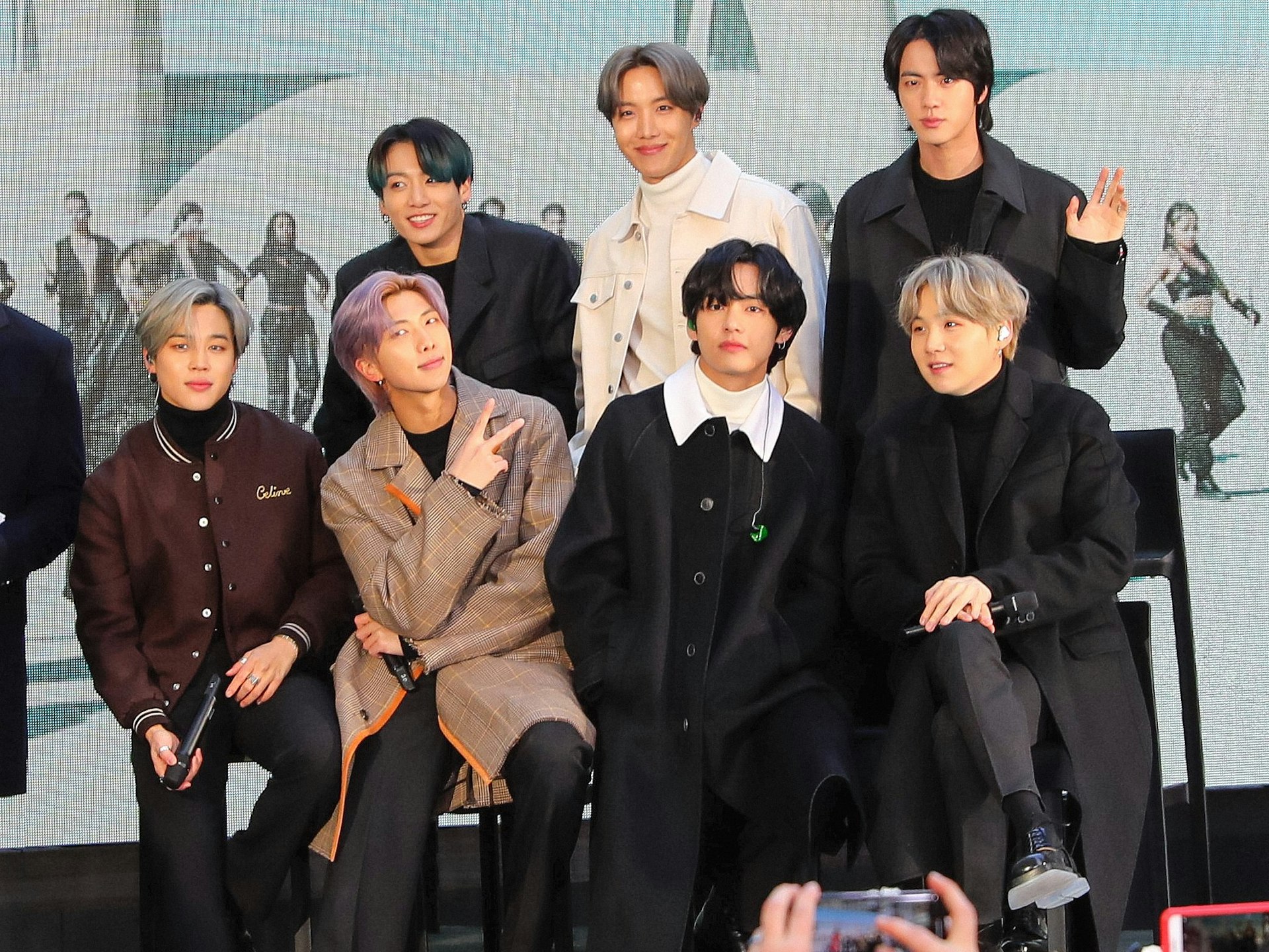 The seven members of K-Pop boy band BTS pose in front of a video screen.