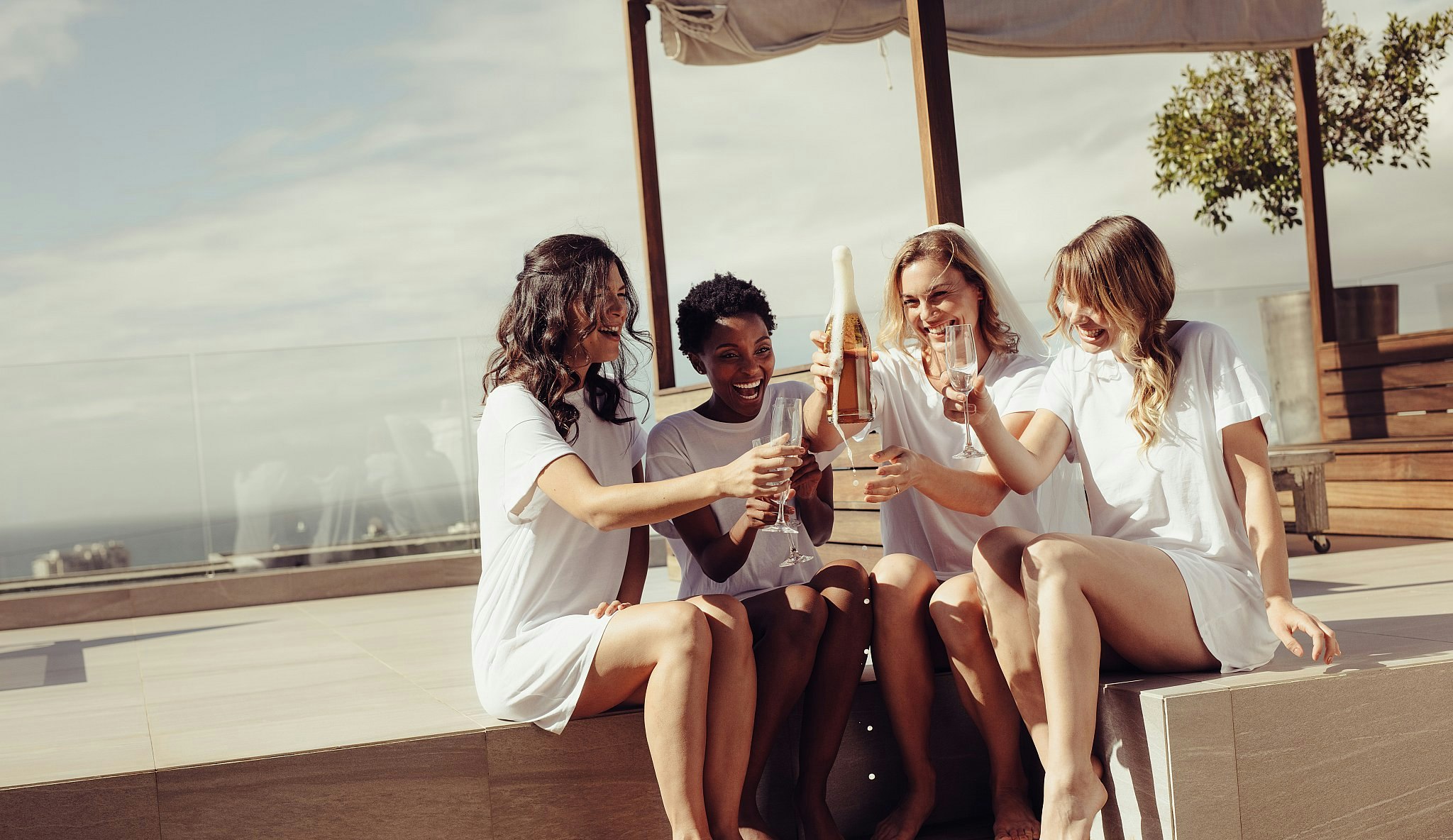 Four laughing young women drinking champagne at a bachelorette party on a rooftop. 