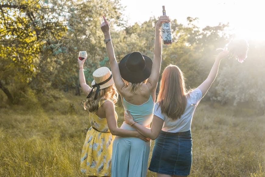 A trio of women with their backs facing the camera stand in the middle of a field, facing the sun. One woman is raising a wine glass, another is raising a wine bottle and another is holding a bouquet of flowers; bachelorette party  