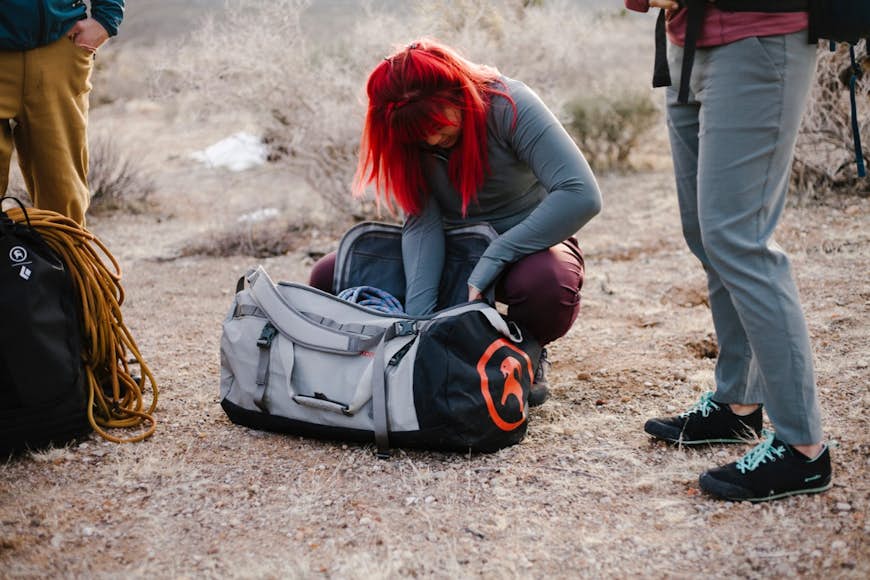 kvinna med knallrött hår, på huk i ett smutsfält och tittar genom en grå Backcountry All-Around duffel, mellan två andra personer i byxor och bredvid en ryggsäck med rep på toppen