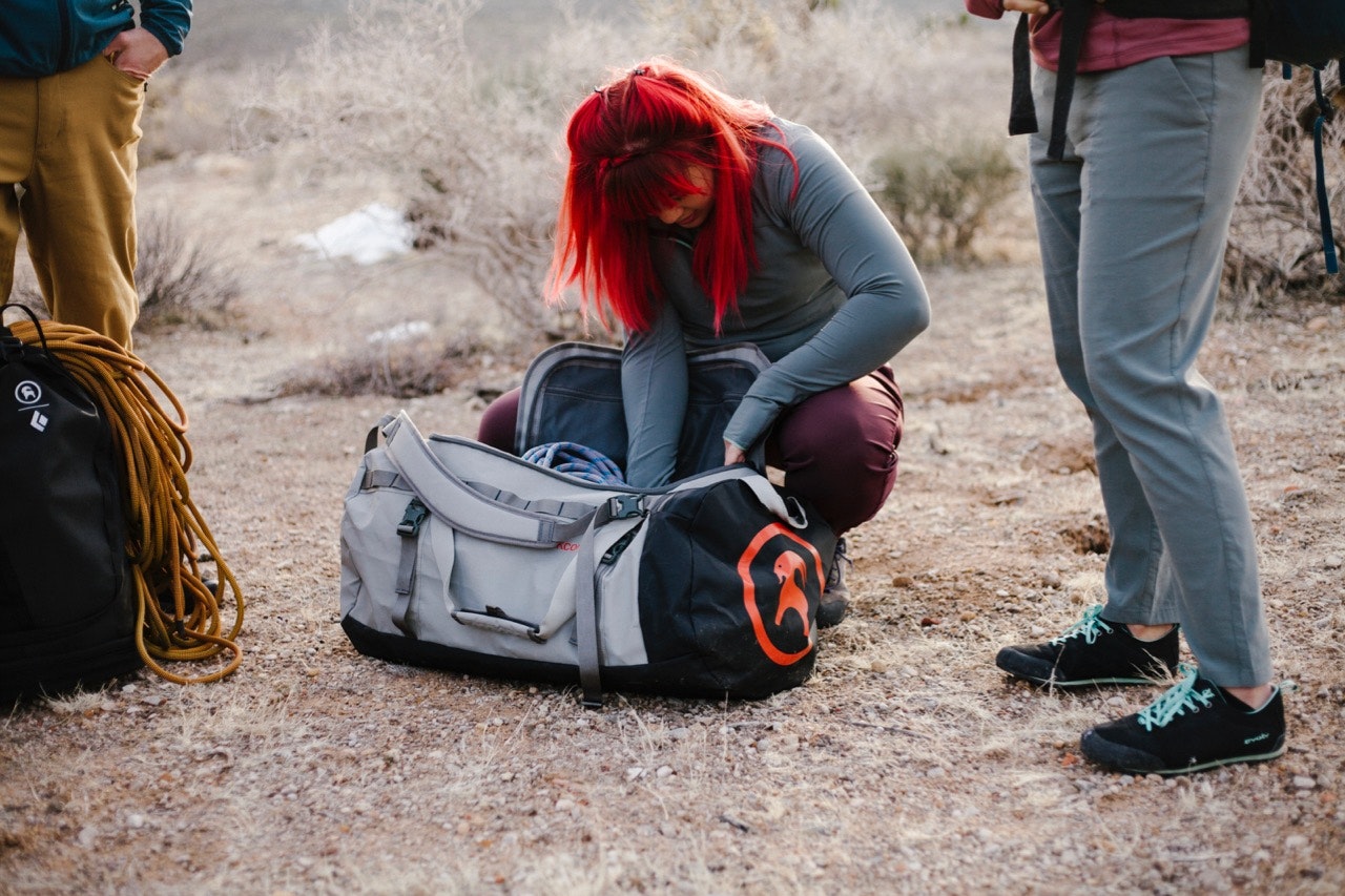 woman with bright red hair, squatting in a dirt field and looking through a gray Backcountry All-Around duffel, between two other people in pants and next to a backpack with rope on top