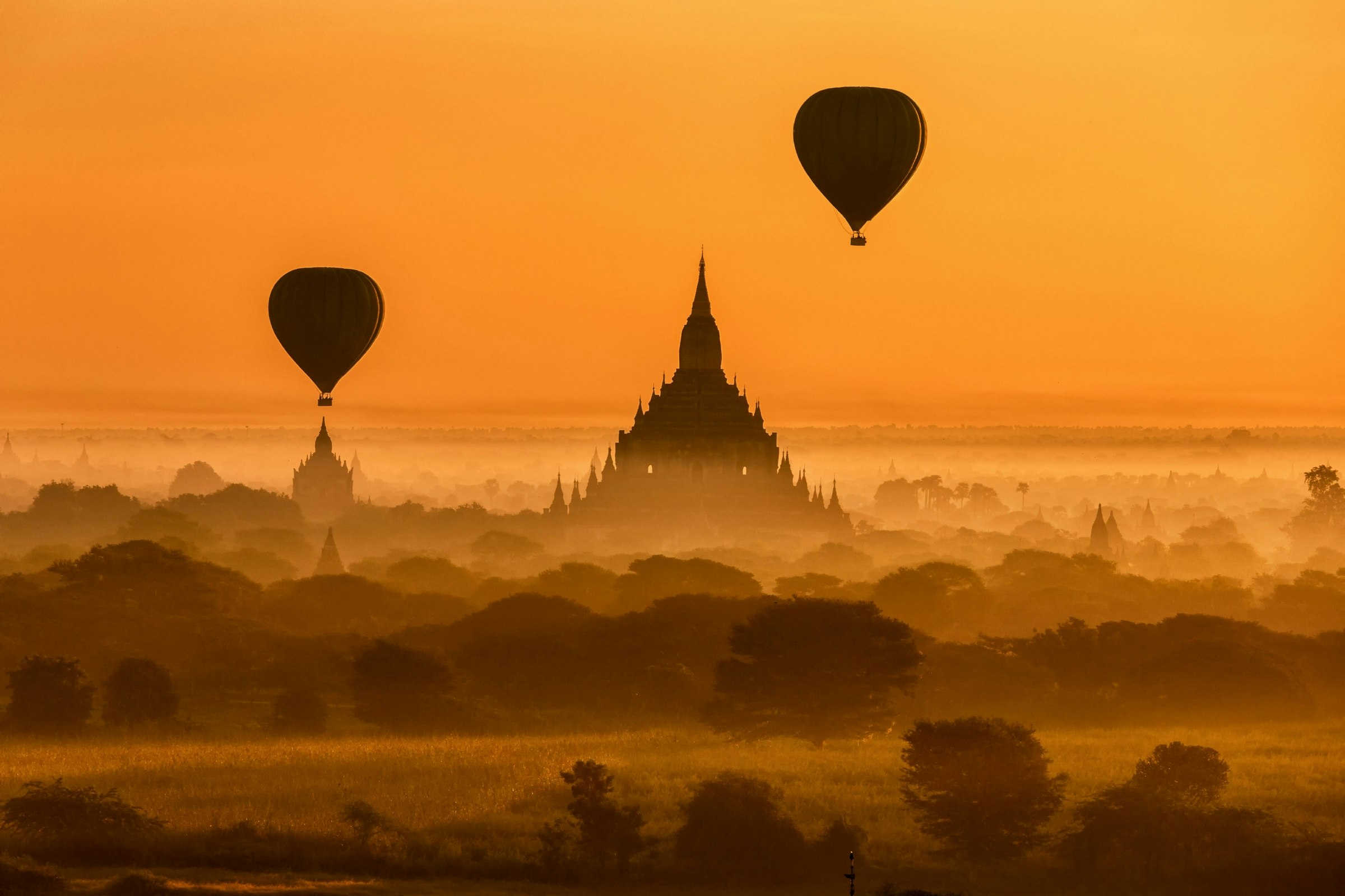 A view overlooking the temples of Bagan at sunset. The ancient temples, surrounded by field and greenery, are silhouetted against the sunset. A couple of hot air balloons also fly overhead, adding to the scenic view.