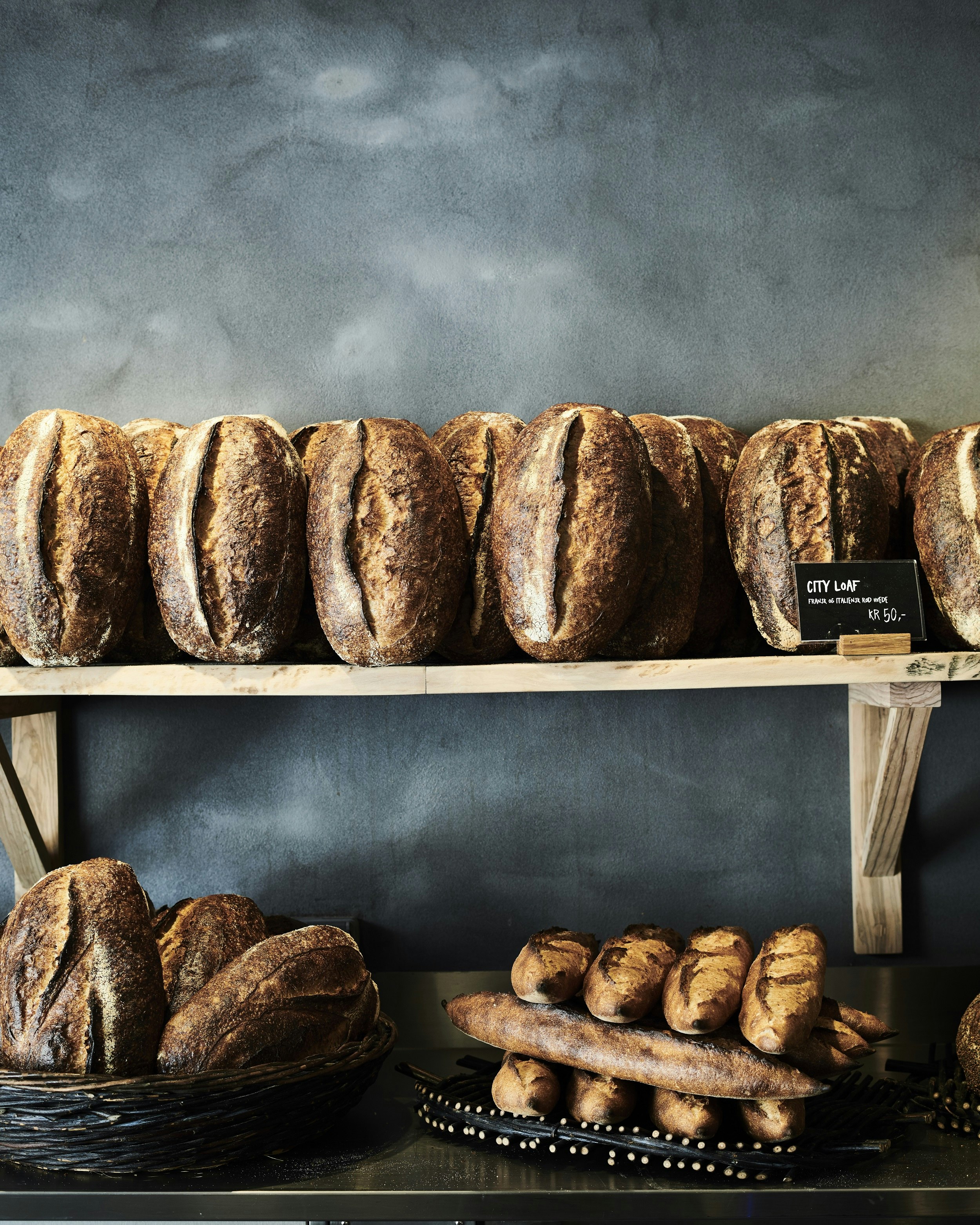 Loaves of freshly-baked bread sitting on wooden shelves; in the background is a dark grey wall. 