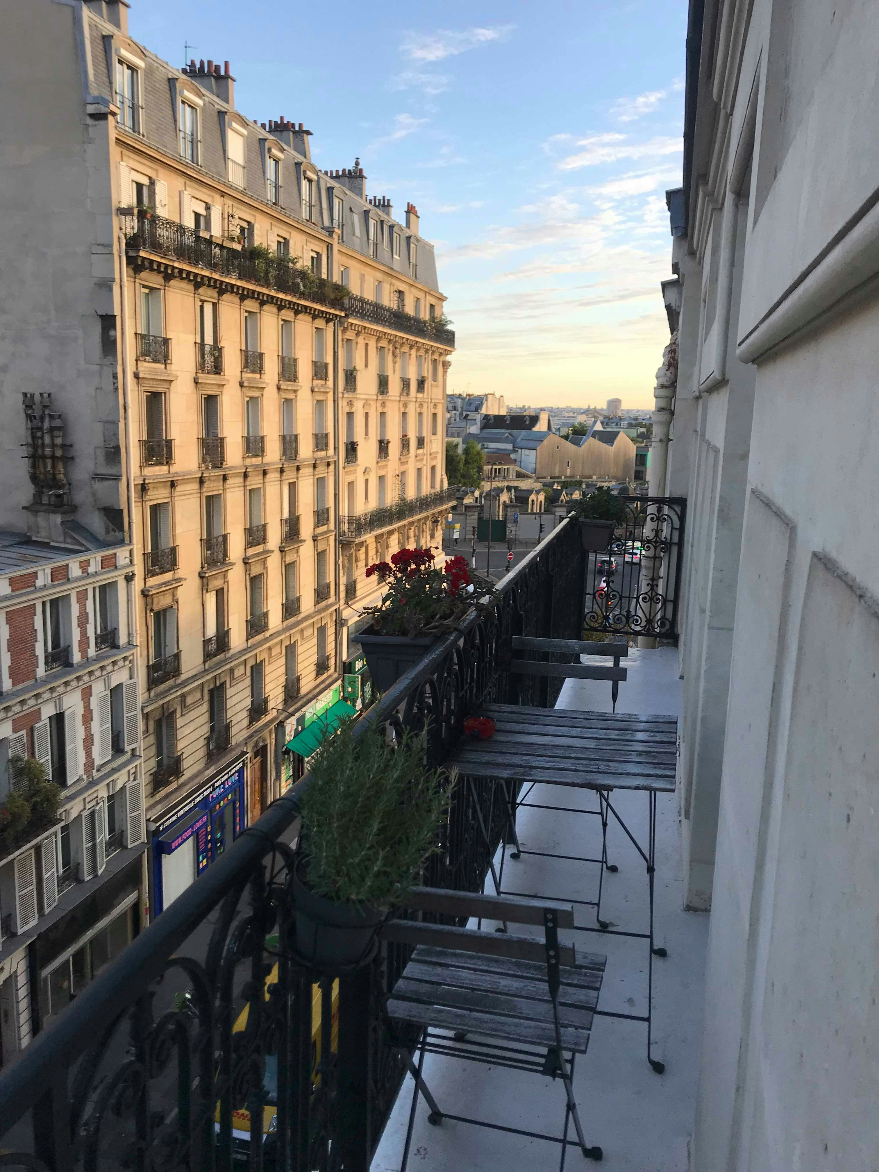 A small table and 2 chairs on a narrow balcony. In the background, the sun lights up a large building