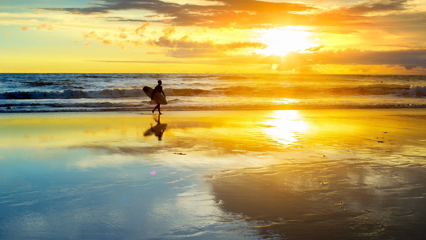Silhouette of a surfer walking on a sandy beach with a surfboard during sunset.