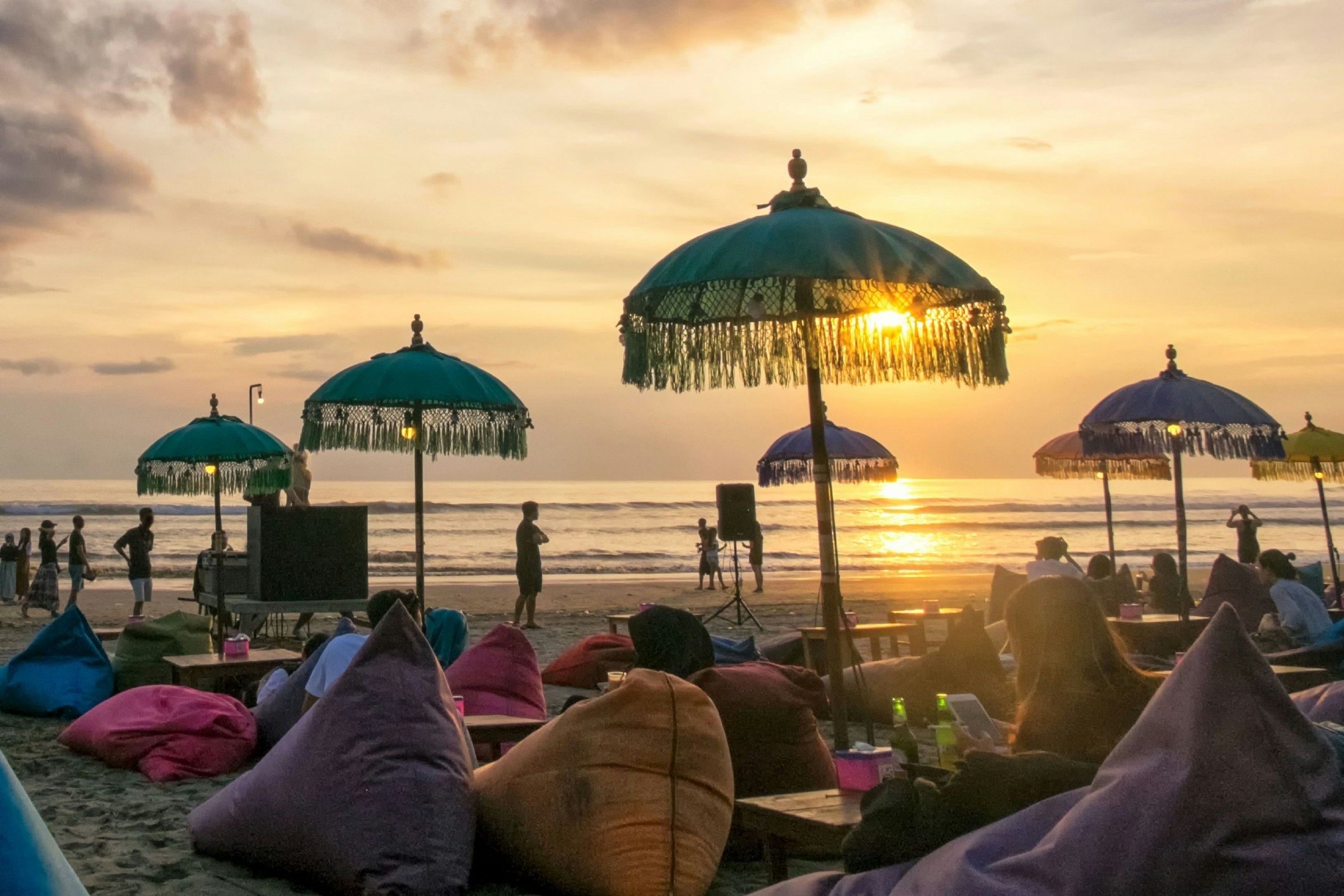 Travellers sit on beanbags scattered on the sands of Seminyak Beach in Bali, Indonesia. The beanbags all have small wooden tables next to them and umbrellas overhead. It is evening and the sun is setting over the ocean in the distance.