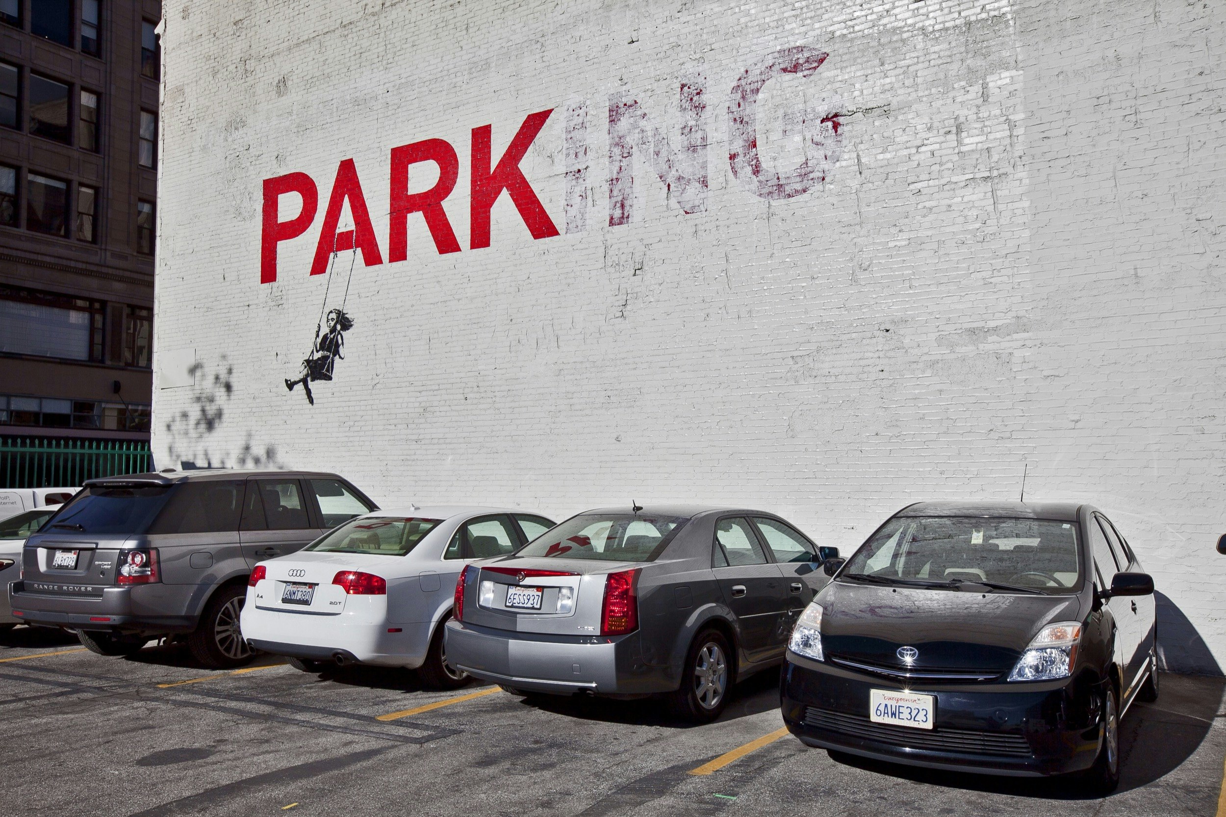 Artwork on the side of a large white wall in Los Angeles depicts a girl on a swing hanging from the A of the word 'Parking'; Banksy in America