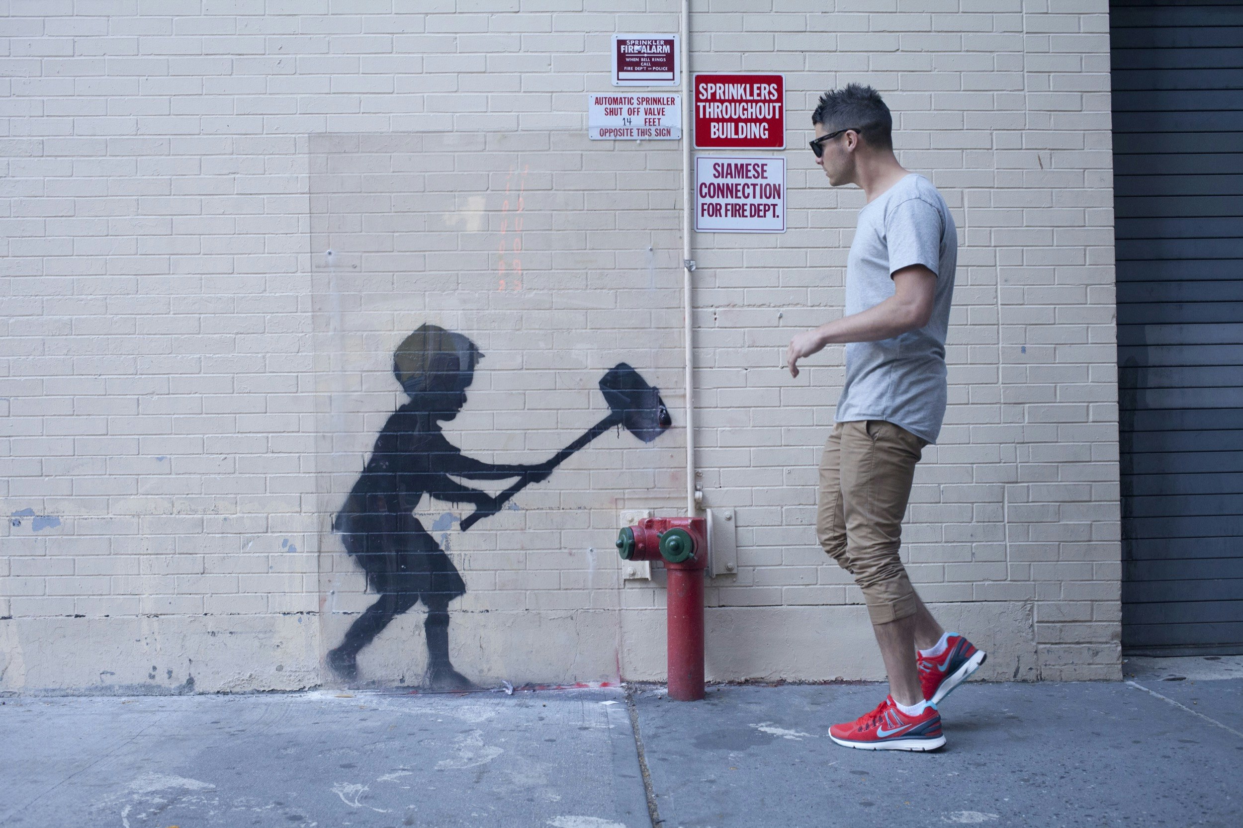 A man walks past a stenciled artwork on a building in New York City, of a boy weilding a hammer; Banksy in America
