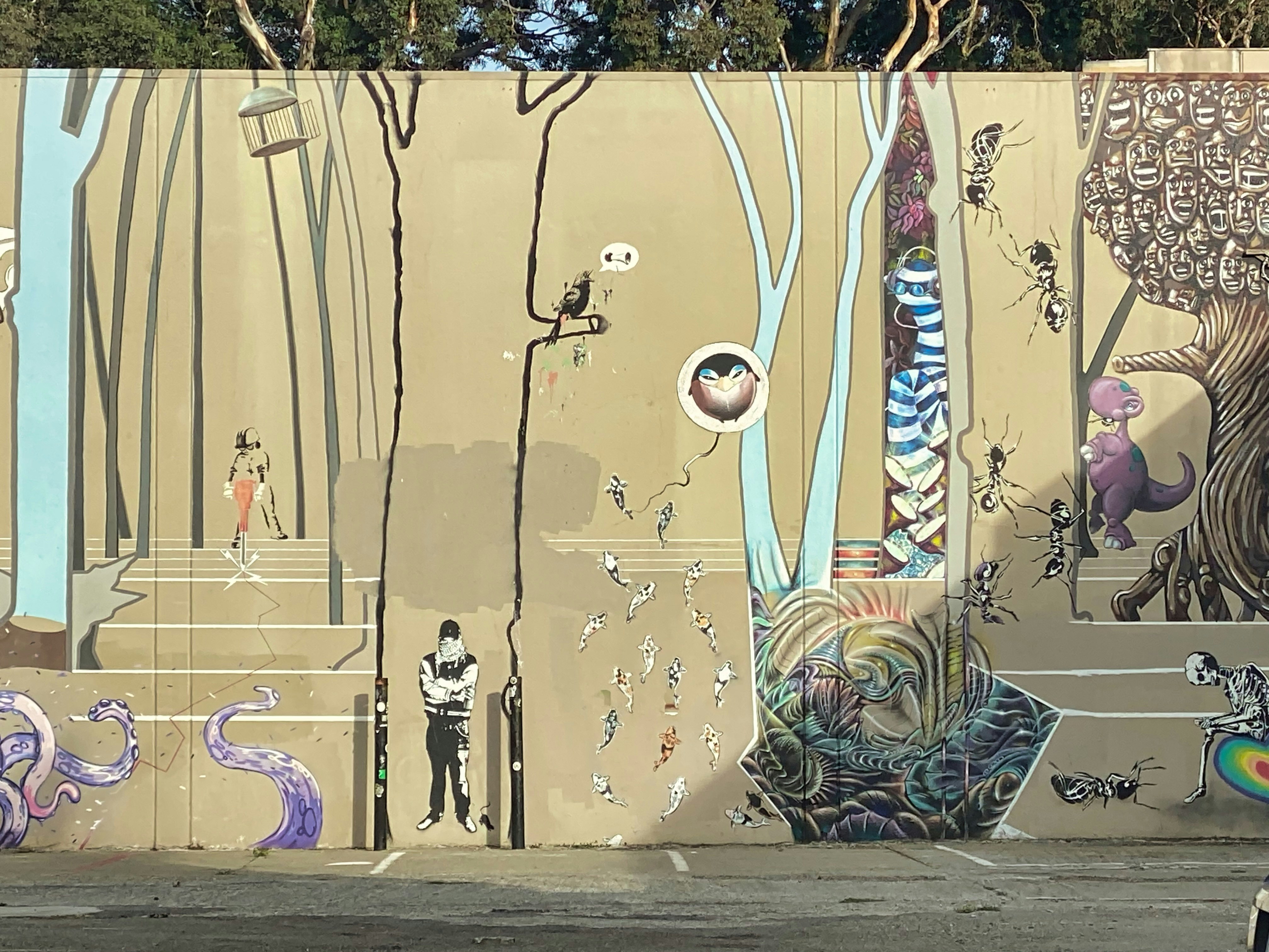 A wall is covered in street art, including a black tree with a bird on a branch over a man with his arms folded